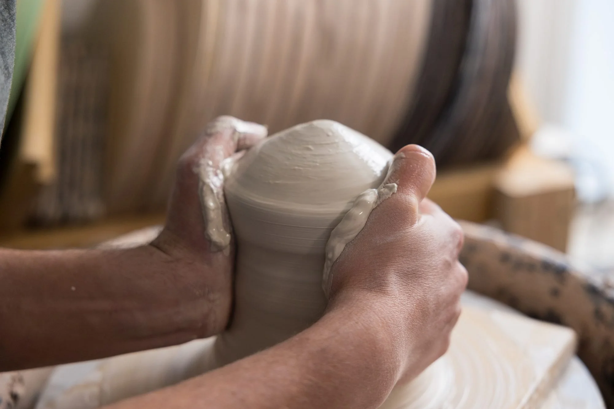 Two hands cupping a mound of clay on a spinning pottery wheel with stacked bats behind