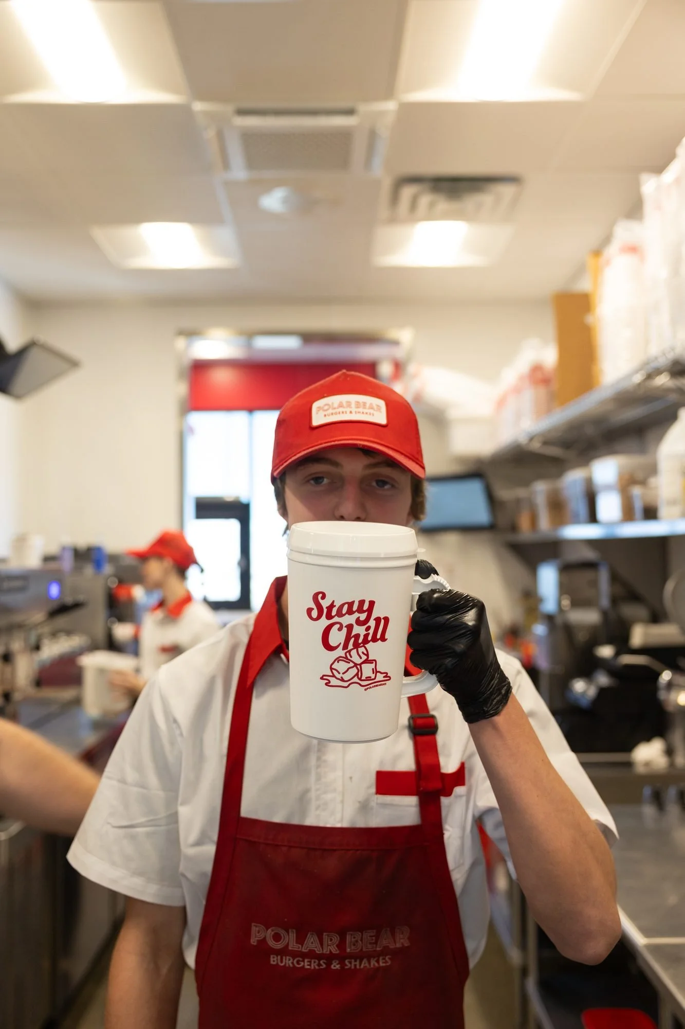 A Polar Bear employee in a red apron and black gloves holding a Stay Chill mug in a busy kitchen