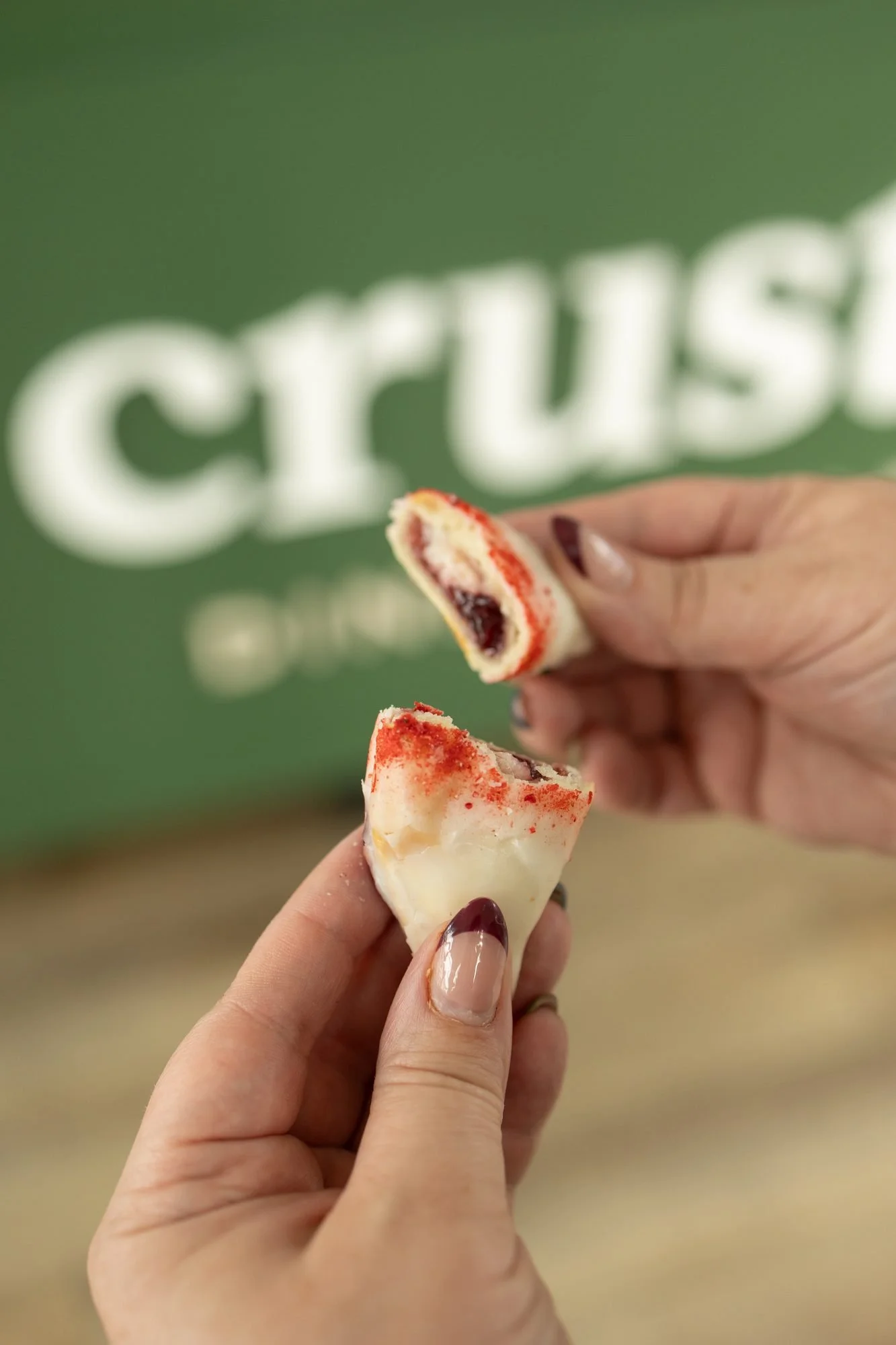 Two hands pulling apart a glazed hand pie with red jam filling in front of a green Crust Club sign