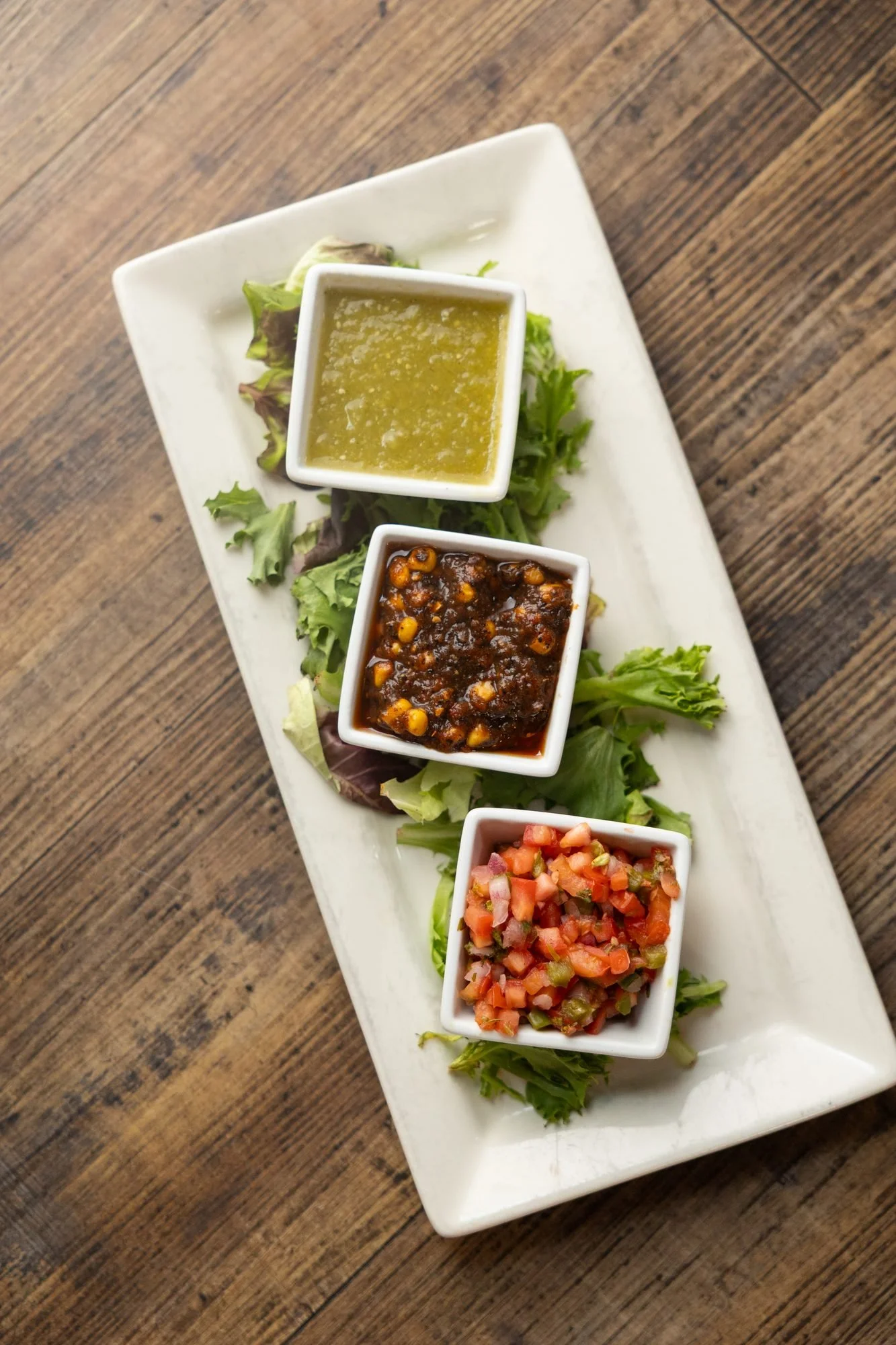 A long white plate with three square dishes of salsas on greens on a wooden table, seen from above