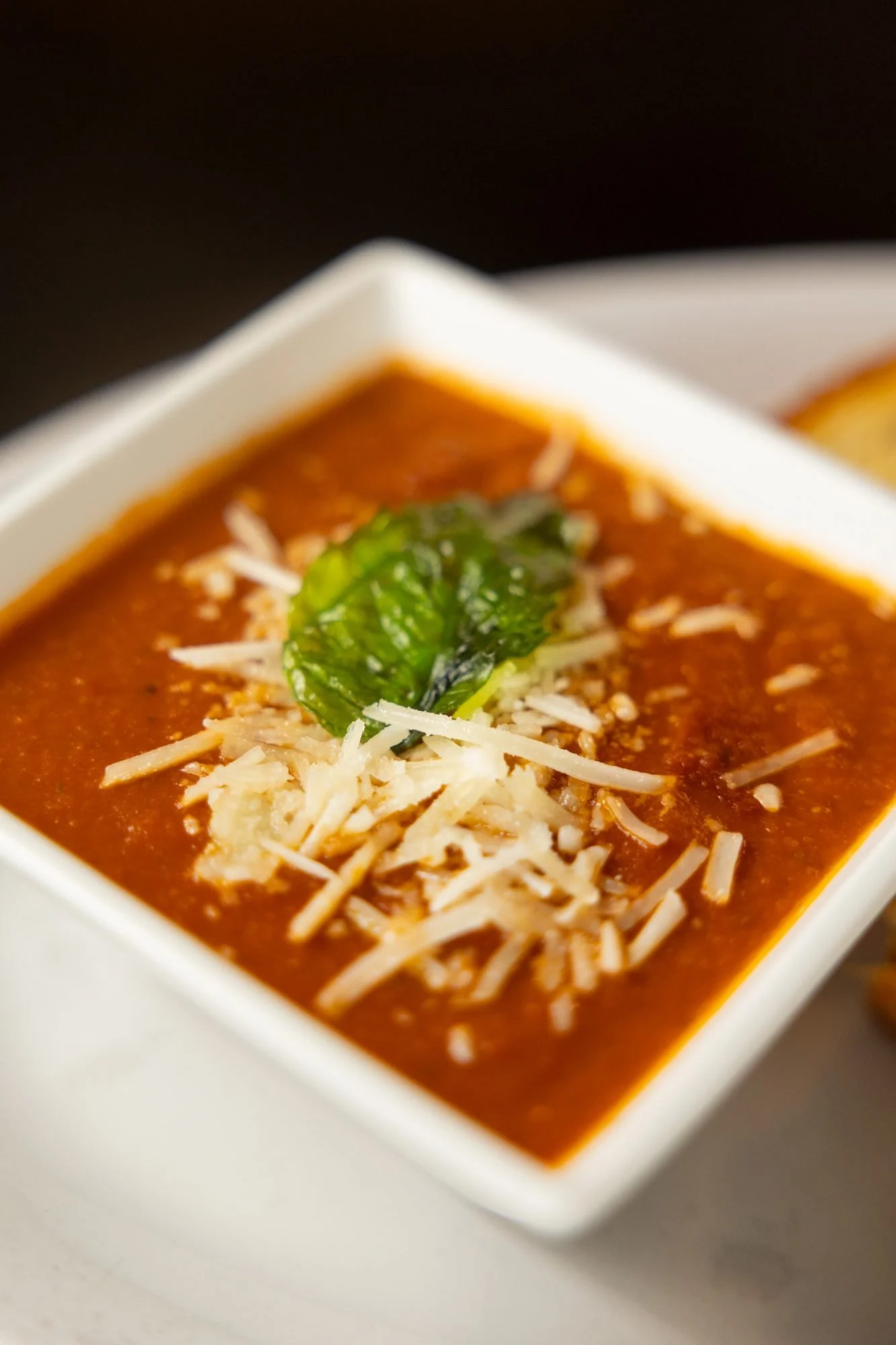 Close view of tomato soup topped with shredded parmesan and a basil leaf in a square white bowl