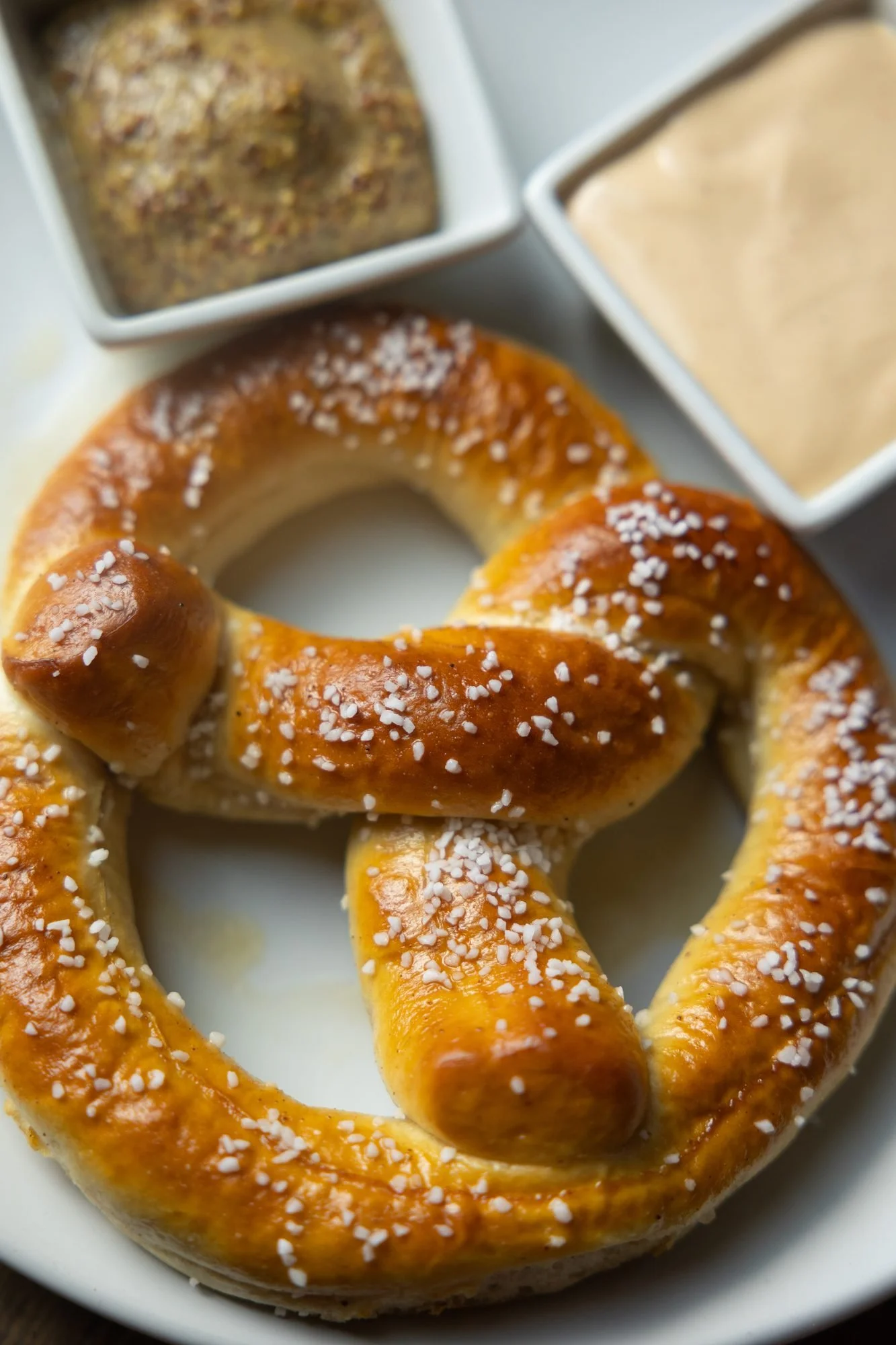 Close view of a salted soft pretzel on a white plate with dishes of mustard and cheese sauce