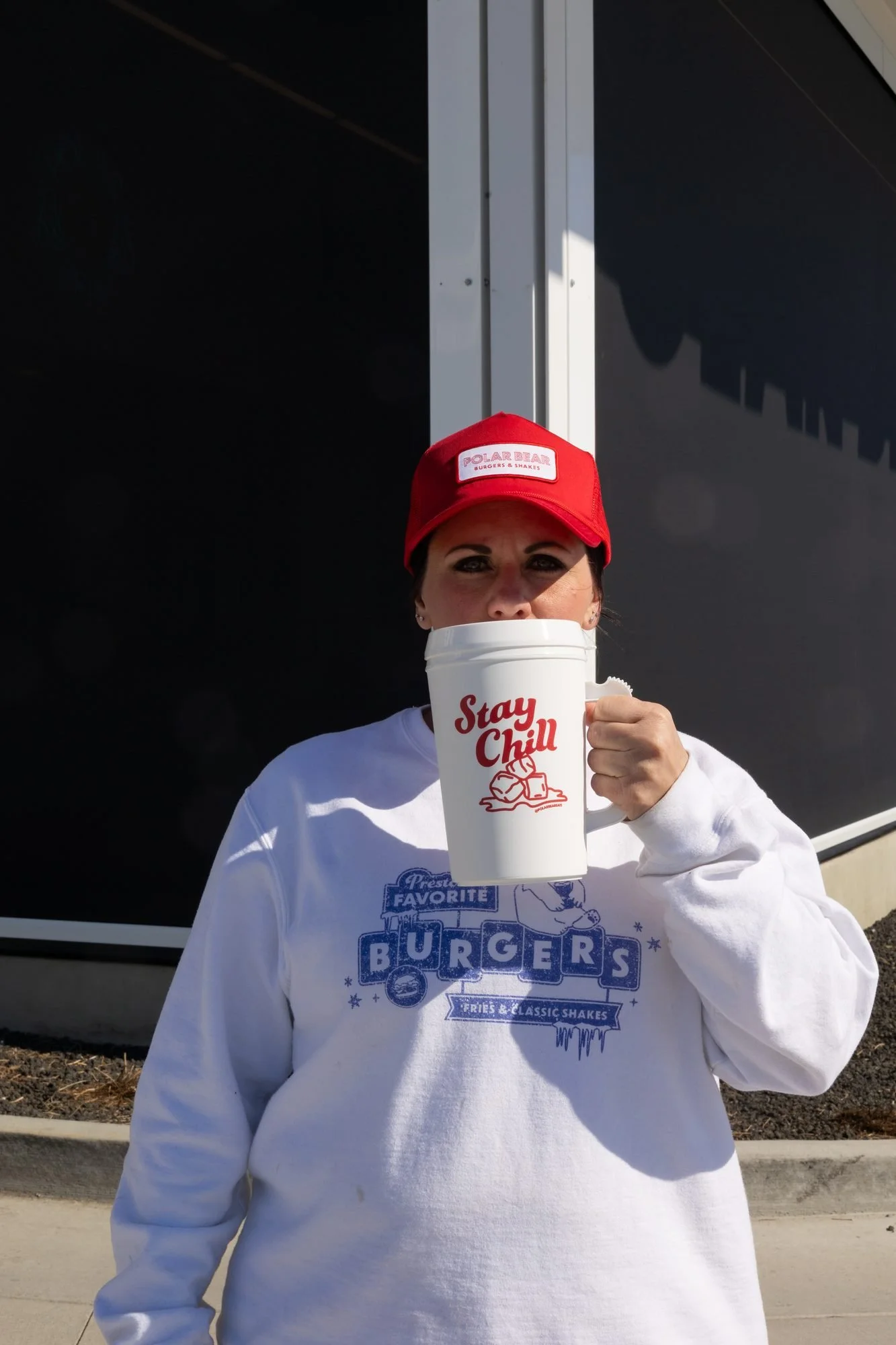 A woman in a red Polar Bear cap and white sweatshirt holding a white Stay Chill mug outside a building