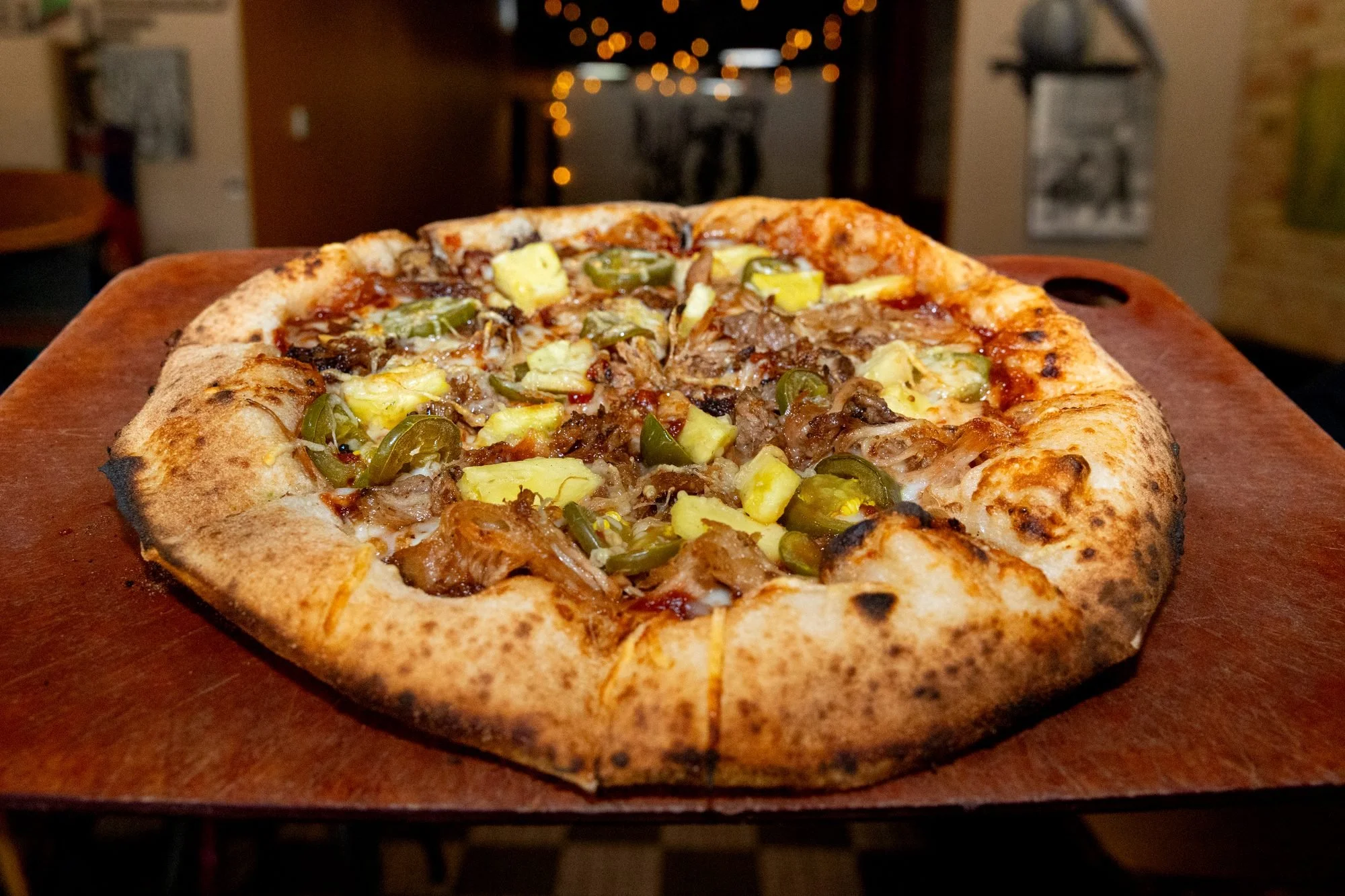 A wood fired pizza with golden crust sits on a wooden board in a dim dining room with warm lights behind
