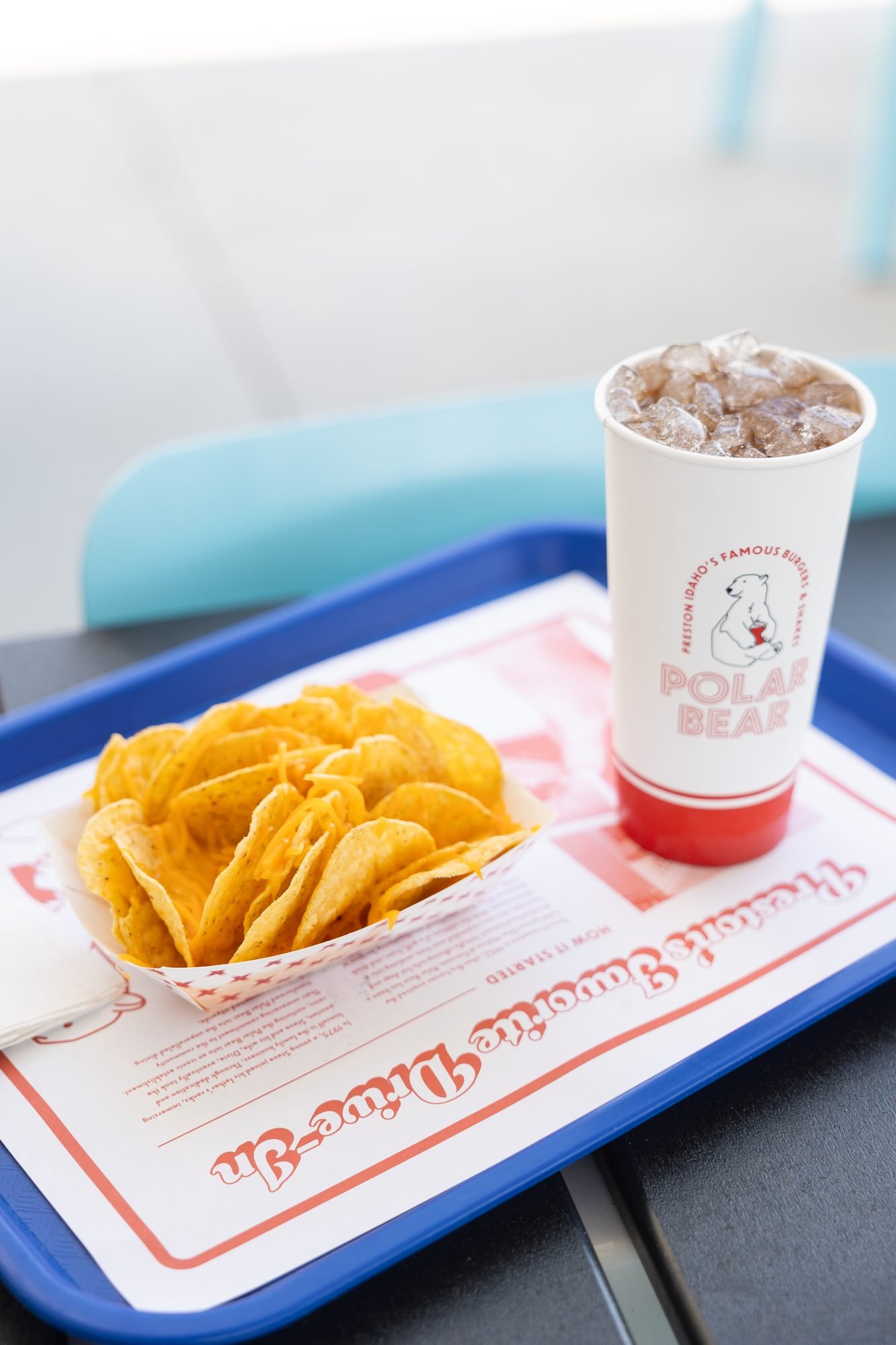 A paper boat of cheesy chips and a soda cup on a blue tray on a table