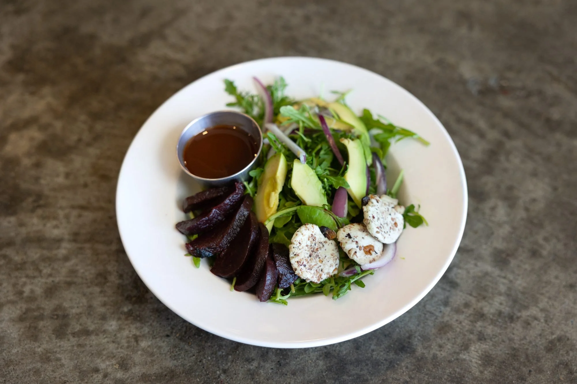A salad with beets, avocado, greens and soft cheese on a white plate, set on a gray concrete table