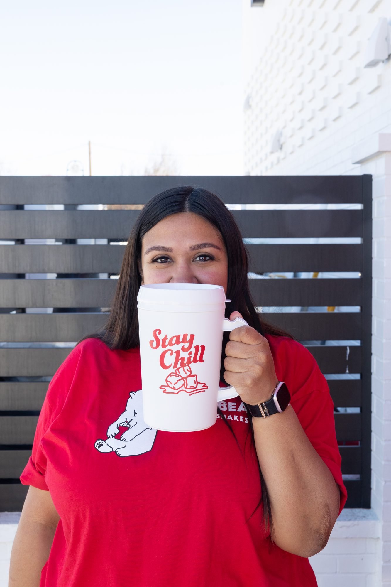A woman in a red Polar Bear shirt holds a Stay Chill mug in front of a dark slatted wall