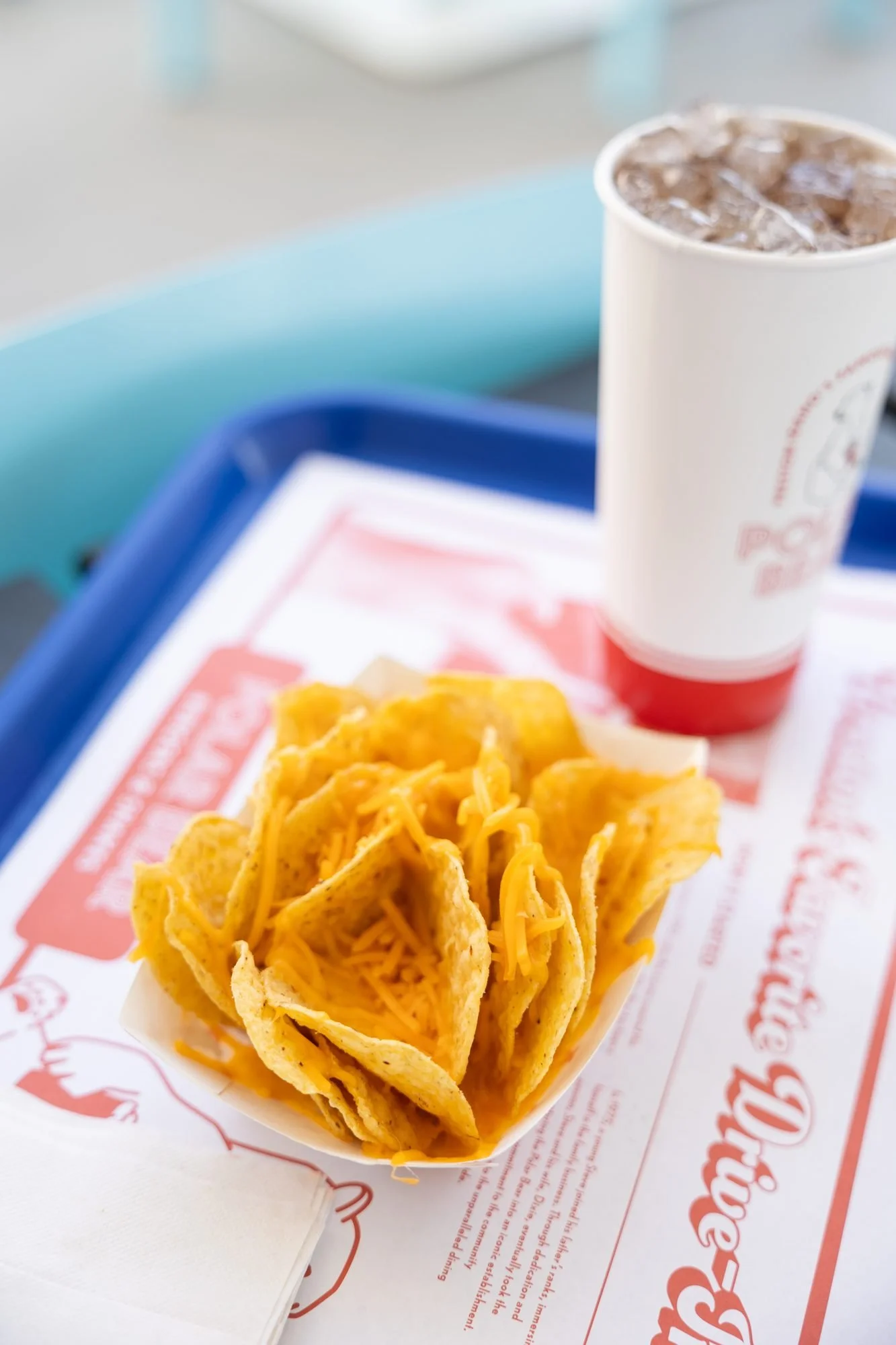 A paper boat of cheesy chips and a cup of soda on a branded tray liner on a blue tray