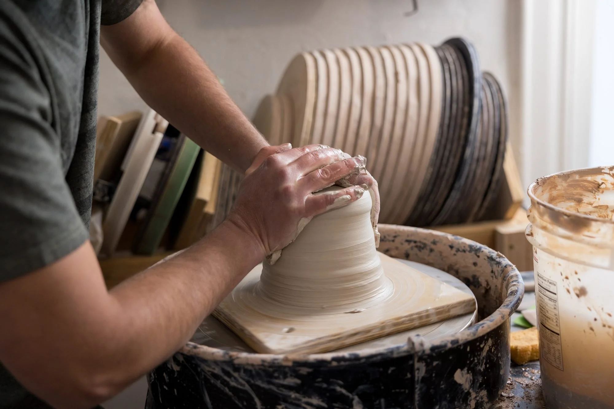 A potter leans over a spinning wheel shaping a mound of clay, with stacked wooden bats behind him