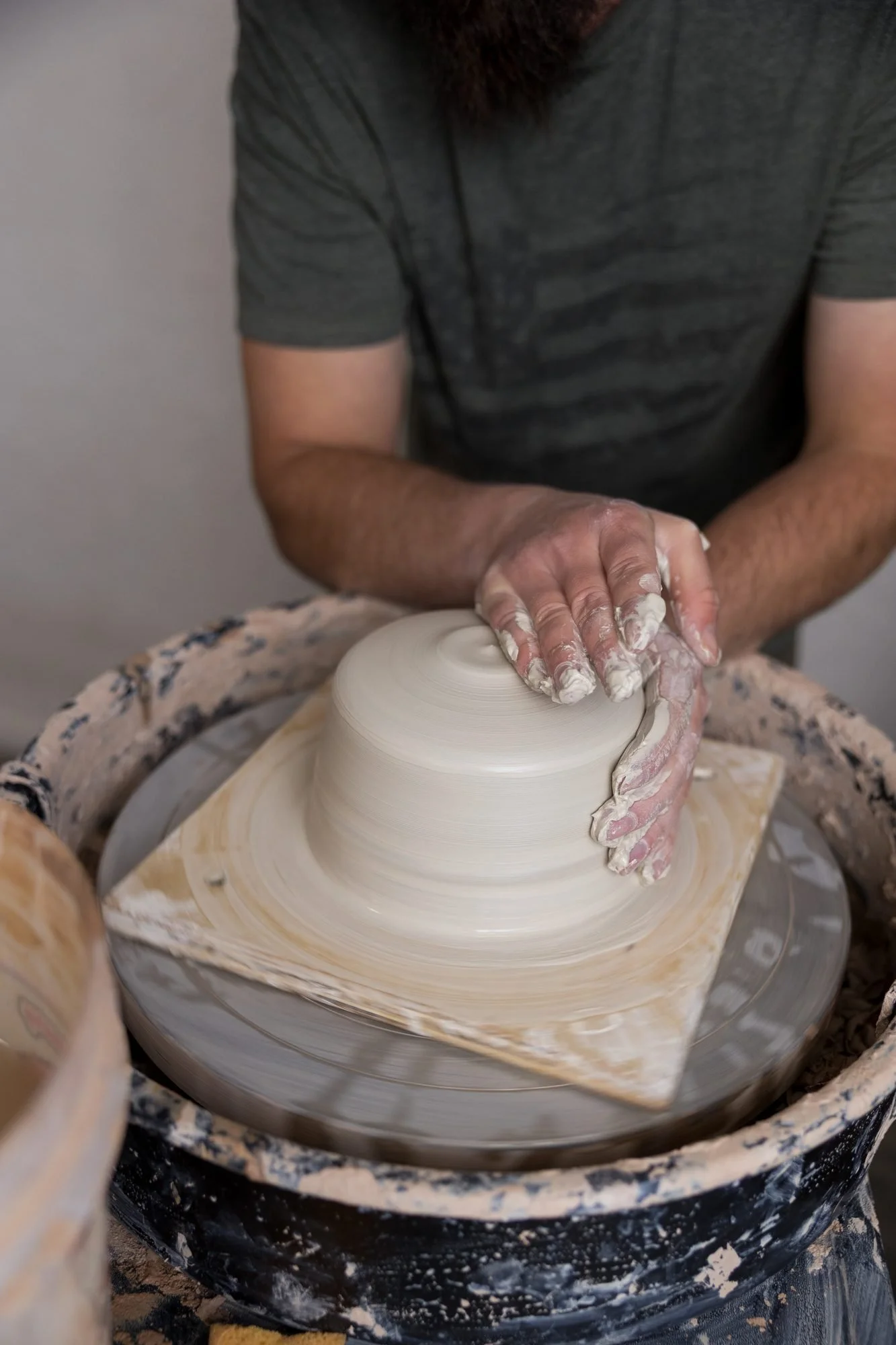 Clay covered hands press down on a wide mound of white clay on a pottery wheel