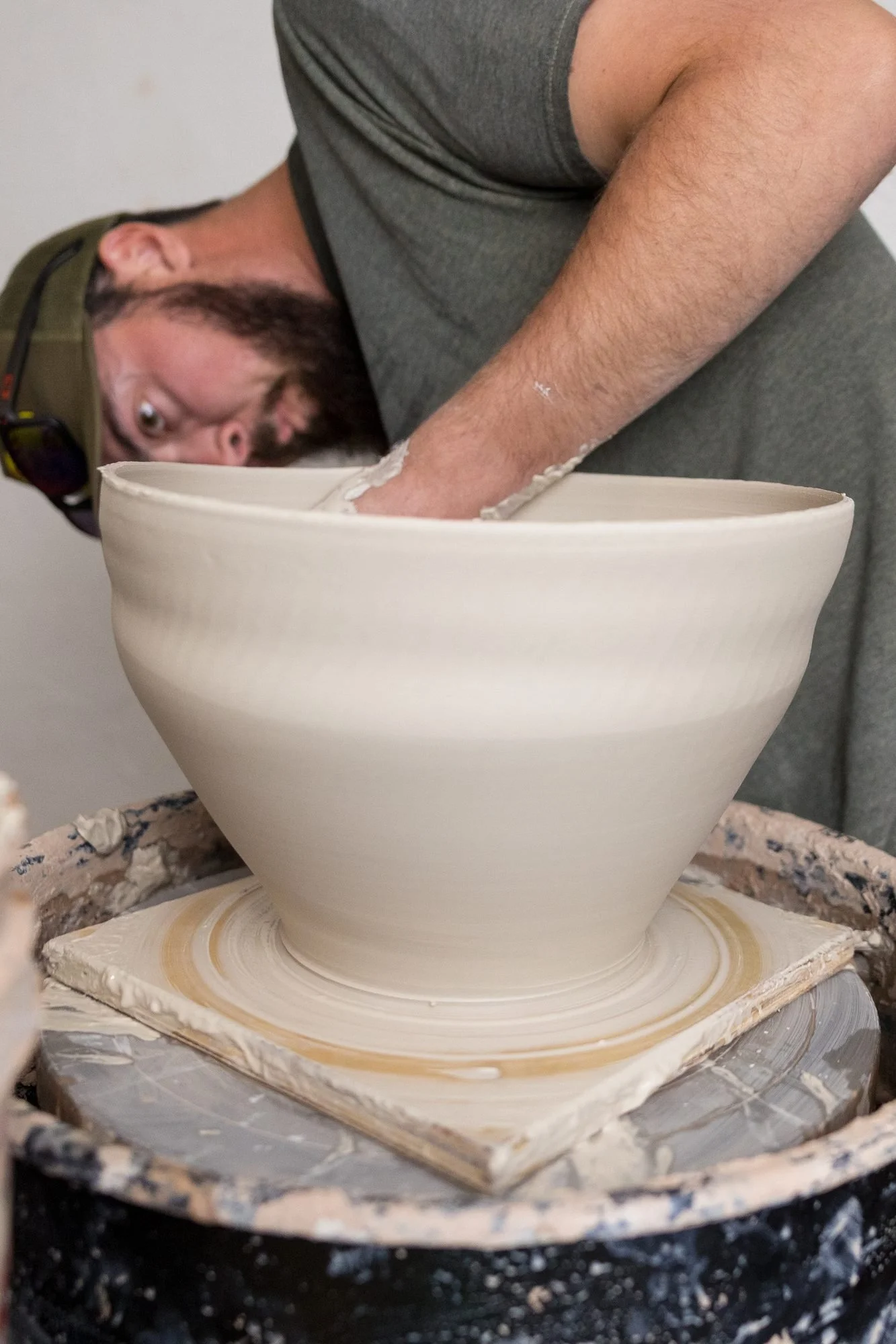 A bearded potter in a cap leans in to check the shape of a large clay bowl on the wheel