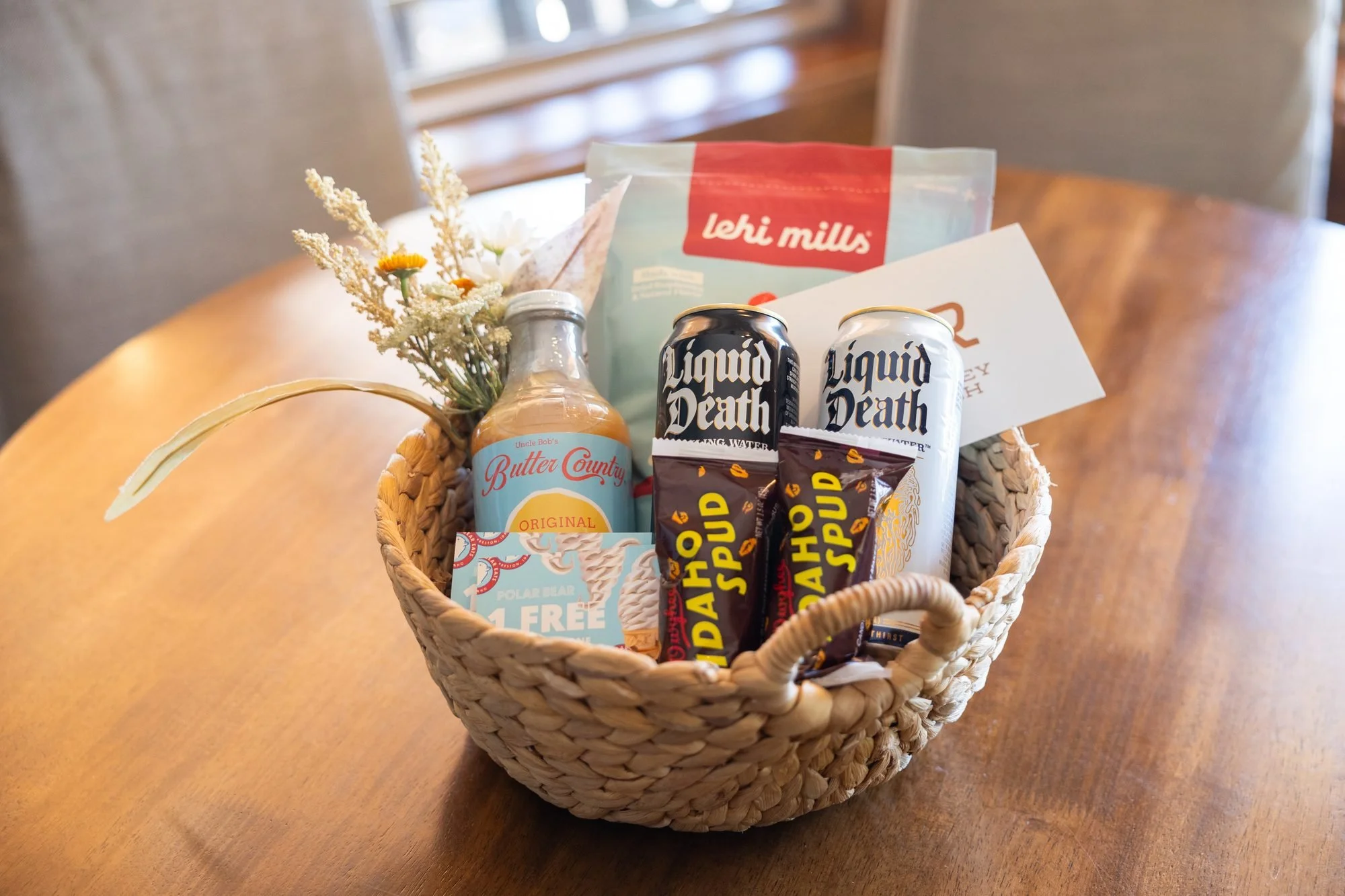 A woven basket of branded drinks, snacks and a small bouquet sits on a round wooden table by a window