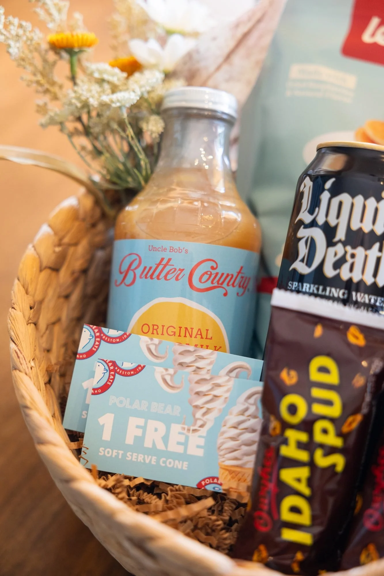 Close view of a woven basket holding a bottle of juice, canned water and potato candy bars with dried flowers