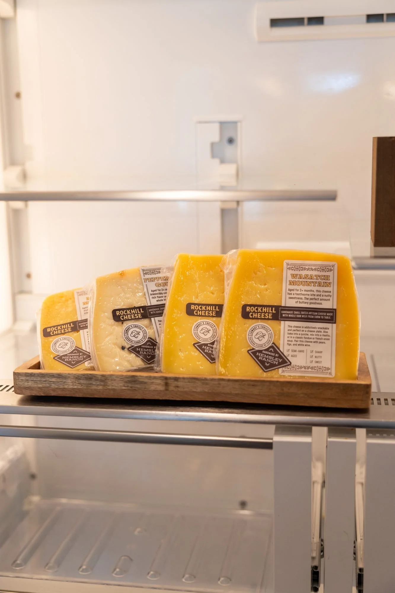 A display case shelf holds wrapped wedges of cheese on a wooden tray