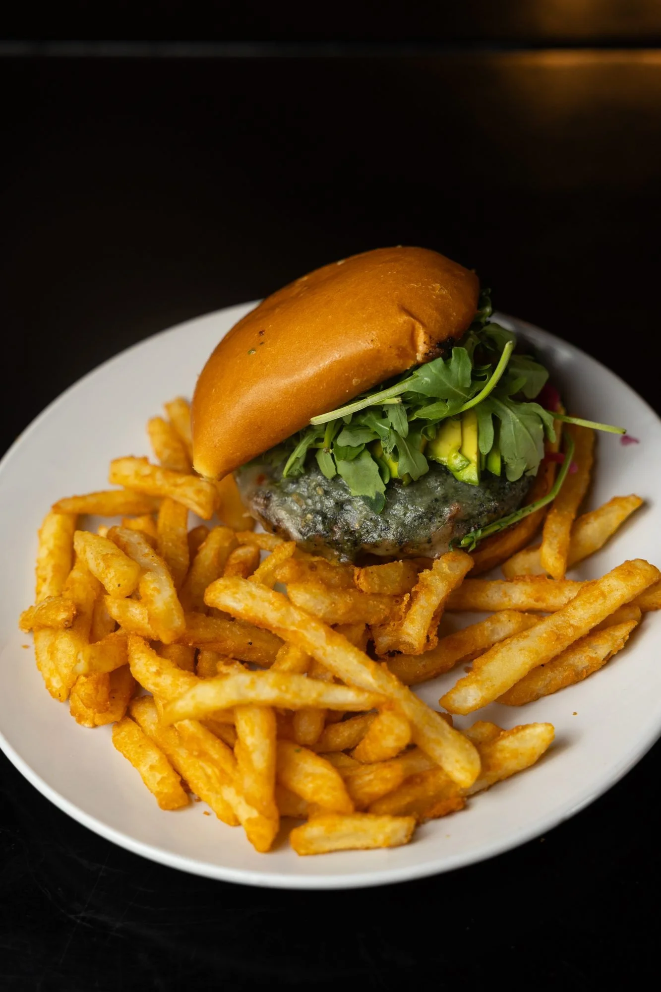 A burger with arugula and avocado next to seasoned fries on a white plate against a dark background