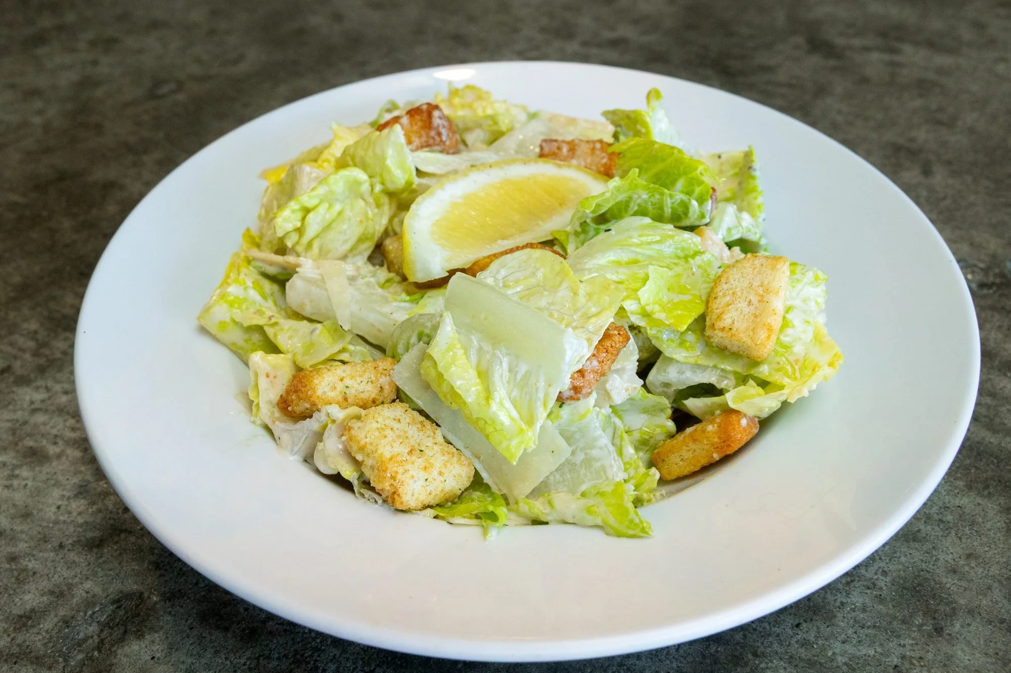 A Caesar salad with croutons and a lemon wedge on a white plate on a gray concrete table