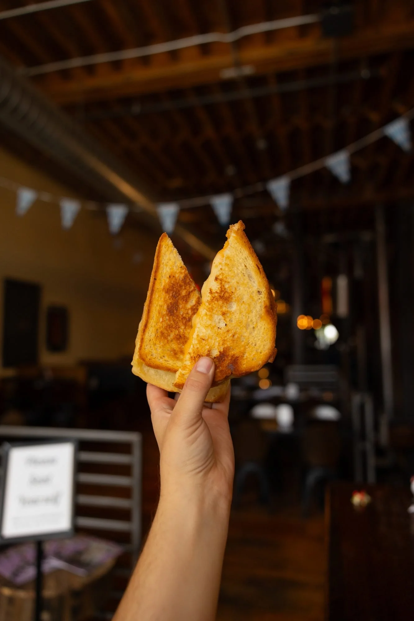 A hand holds up two halves of a grilled cheese sandwich inside a dim restaurant with bunting overhead