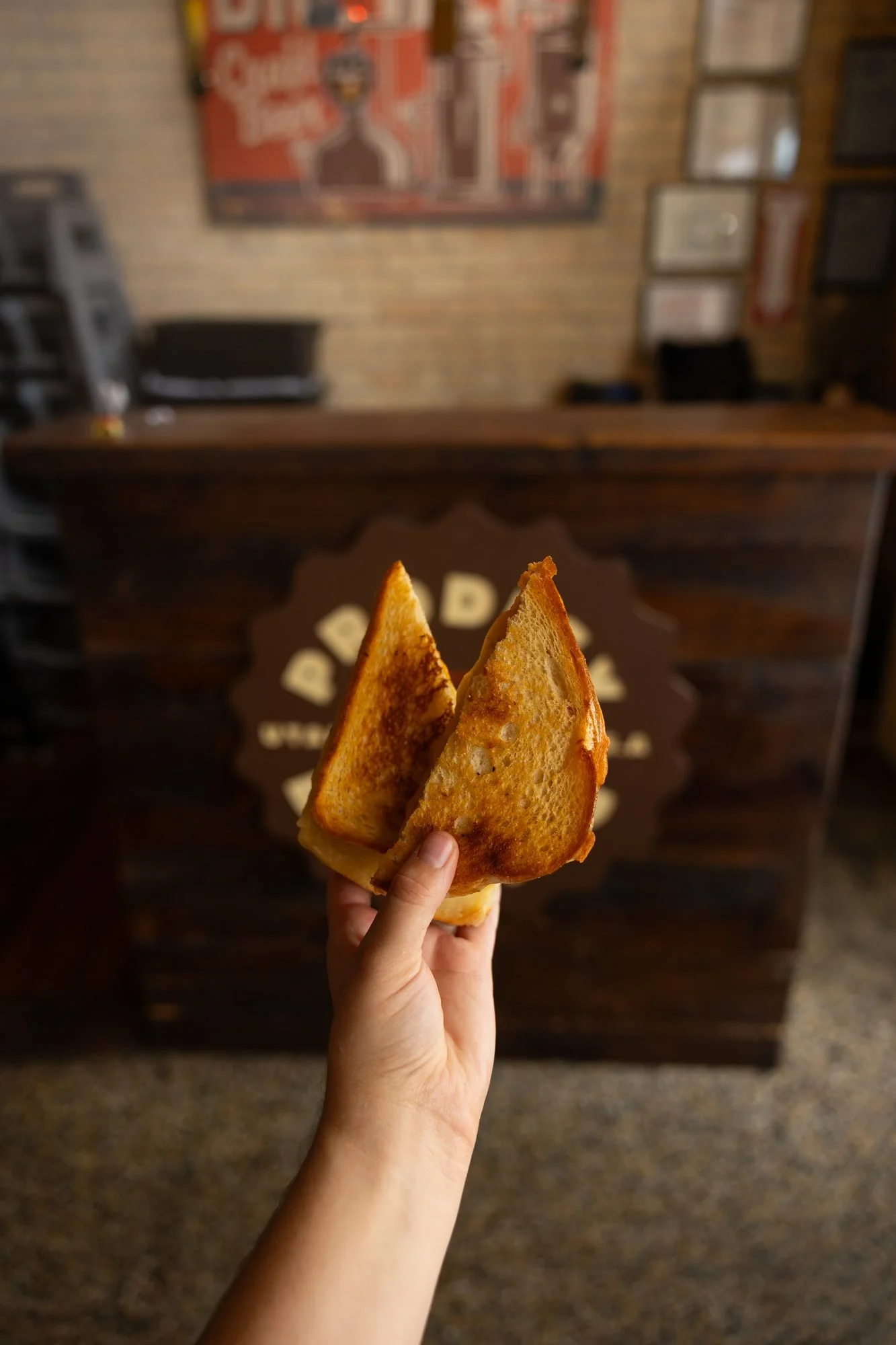 A hand holds two grilled cheese halves in front of a wooden counter with the Prodigy logo