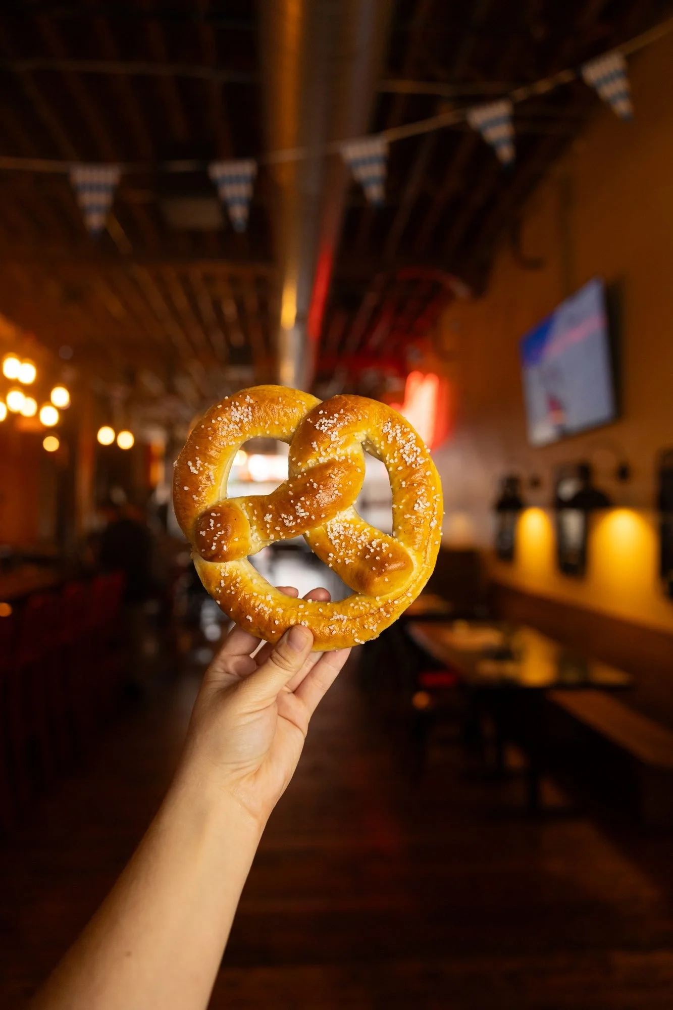 A hand holds up a soft pretzel inside a dim restaurant with bunting and warm lights