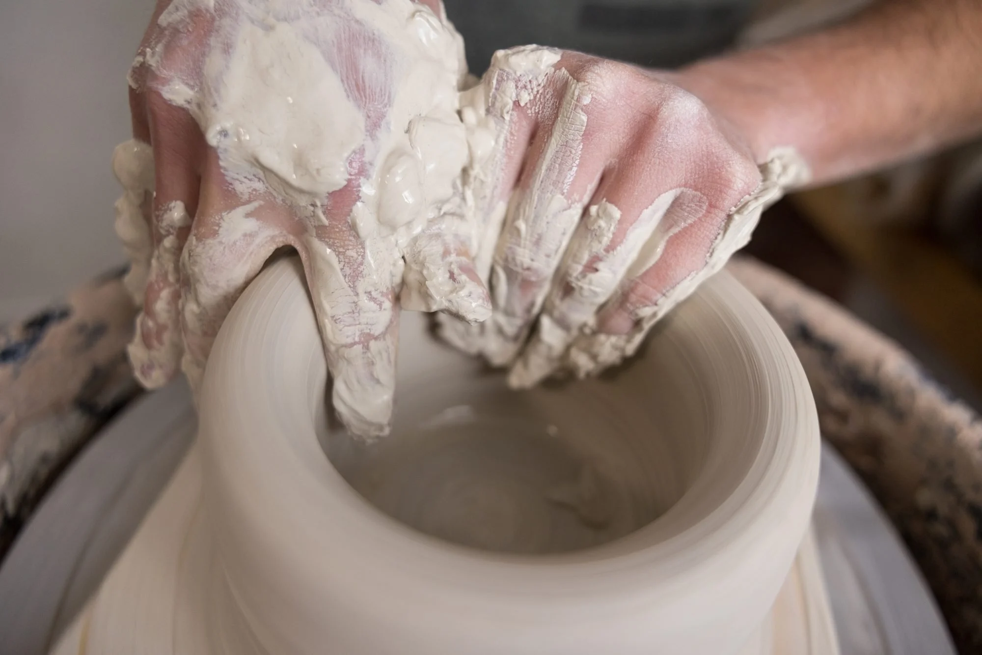 Two clay covered hands shape the rim of a clay bowl on a pottery wheel