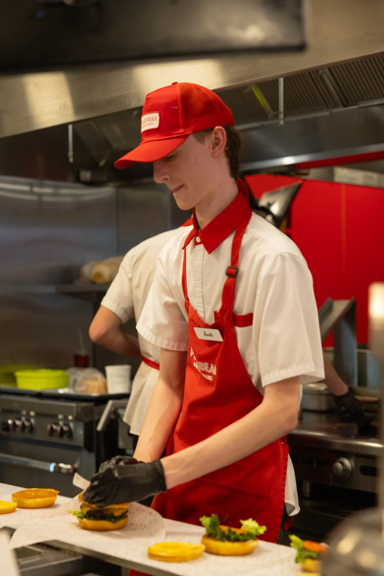 A smiling Polar Bear employee in a red cap and apron assembles burgers on a kitchen line