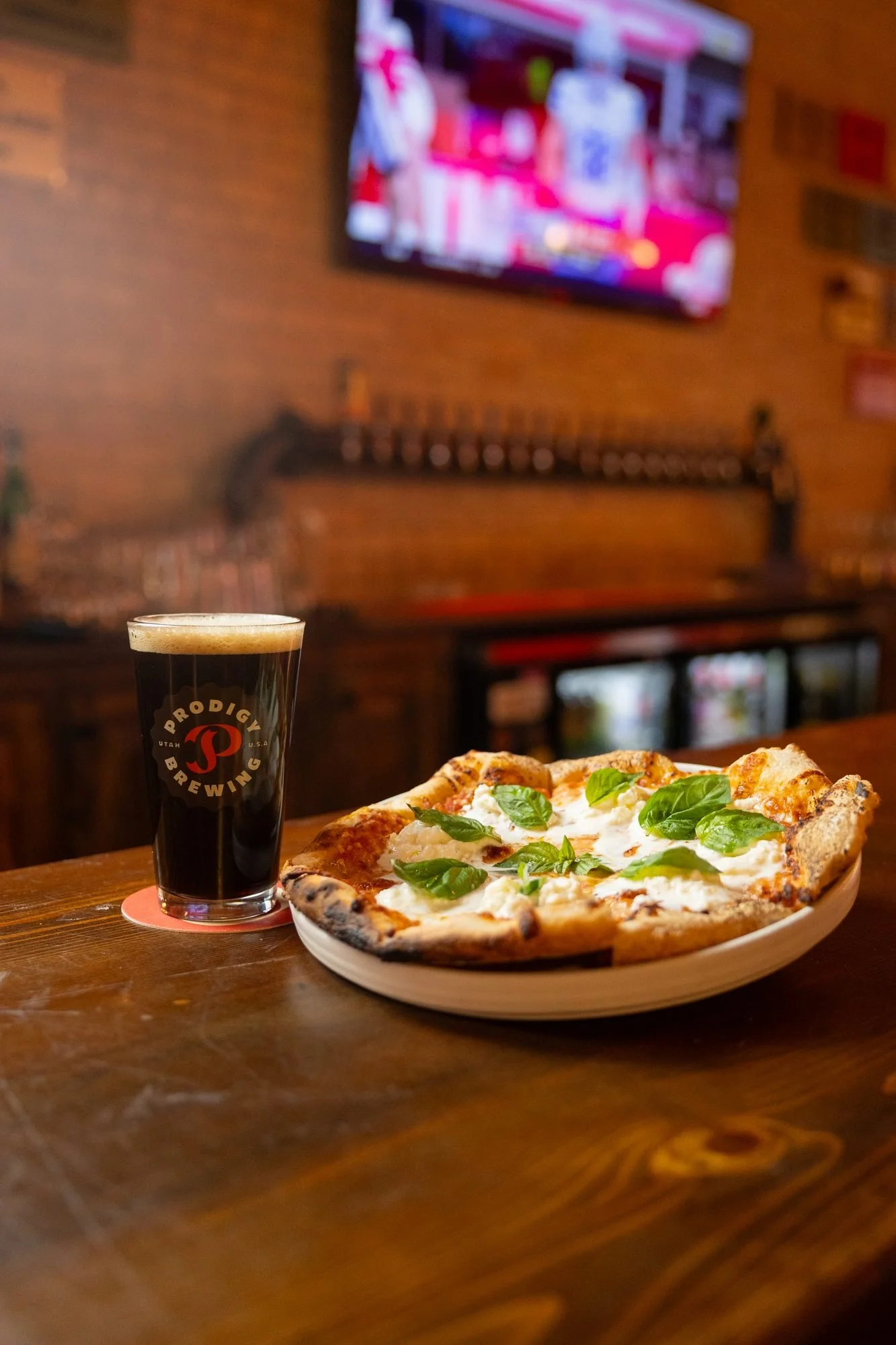 A pint of dark beer beside a pizza with fresh basil on a wooden bar under a television