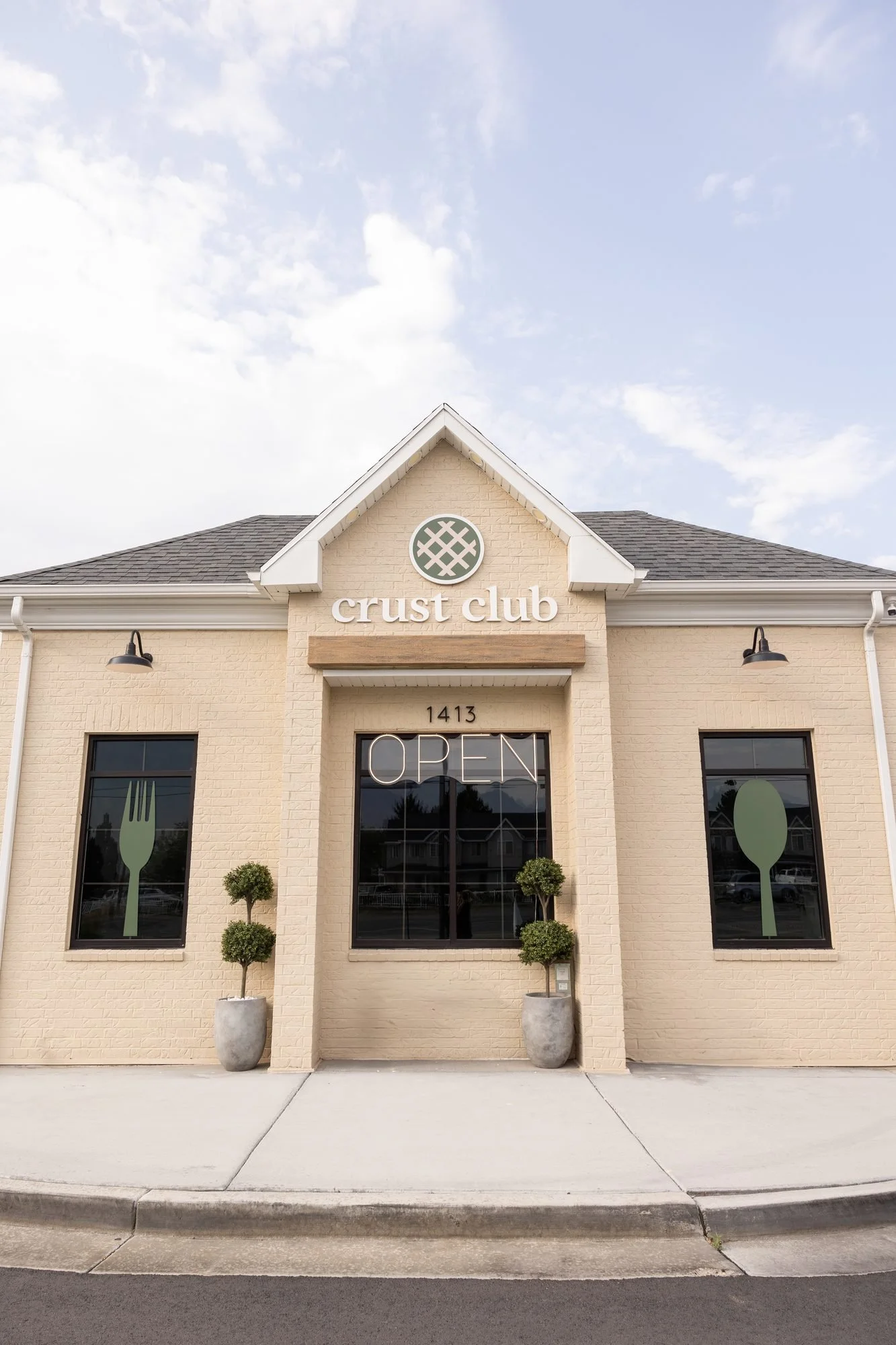 The cream brick front of the Crust Club building with an OPEN sign and potted topiaries by the door