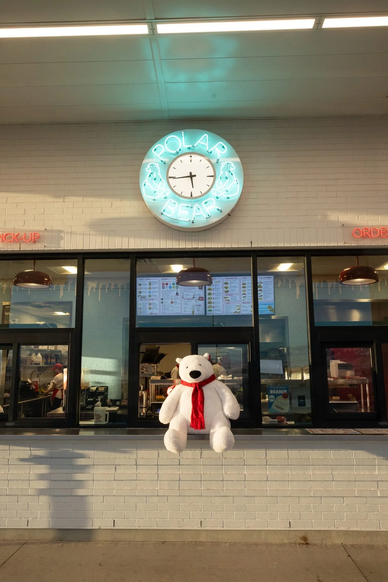 A stuffed polar bear in a red scarf sits outside a Polar Bear order window under a neon clock