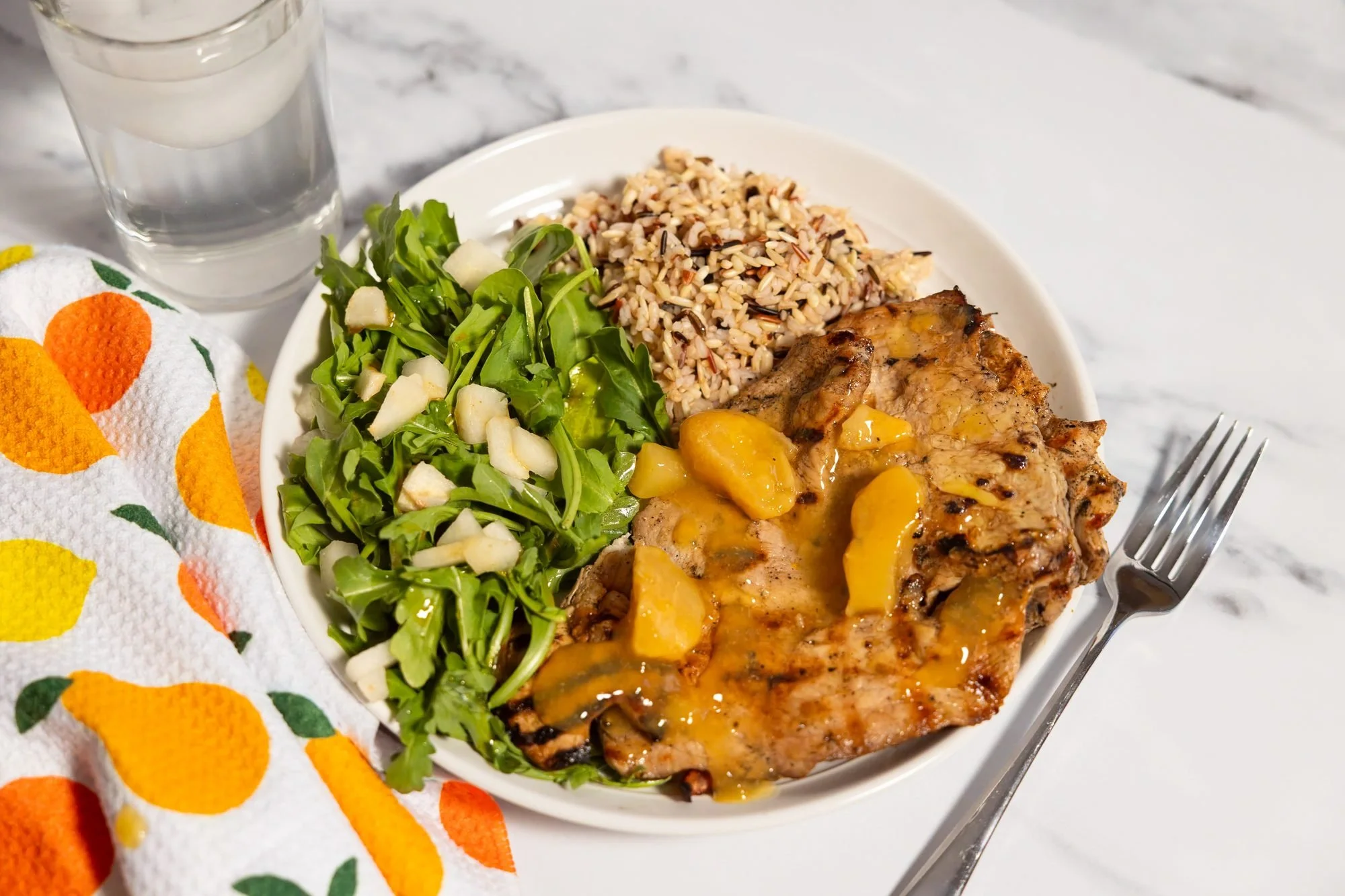 A plate of grilled chicken with peach sauce, arugula salad and wild rice beside a fruit-print towel and water glass