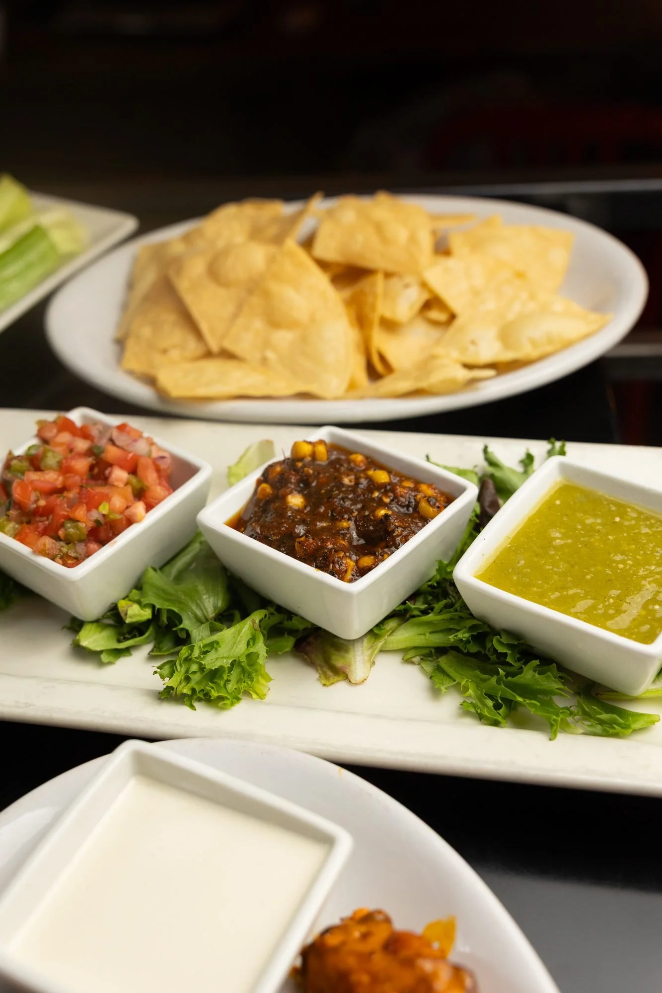 Tortilla chips and three salsas in white dishes on a long plate with a bowl of ranch in front