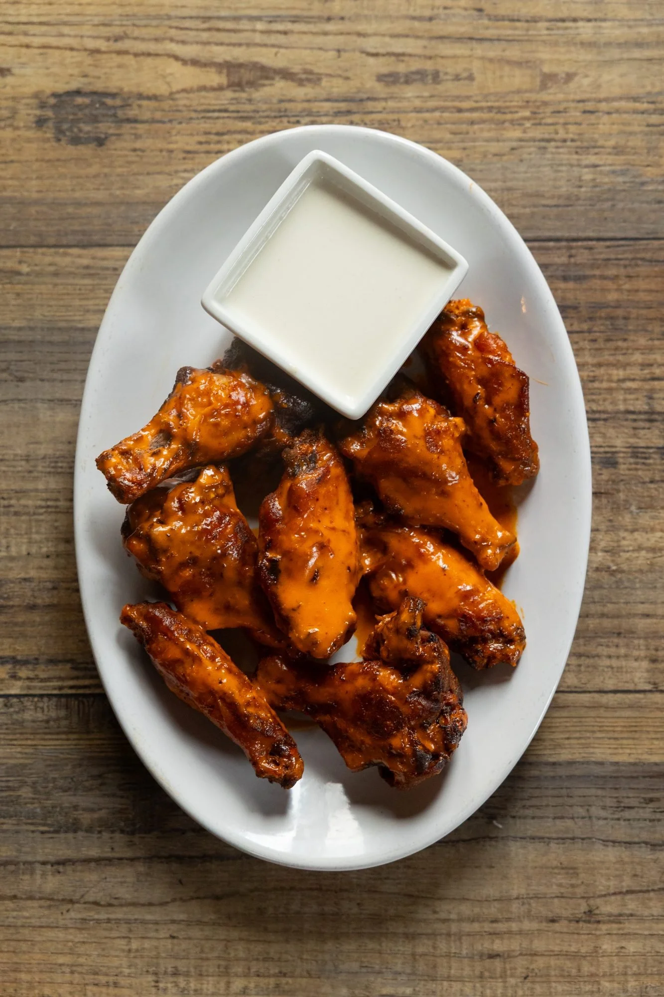 A plate of glazed chicken wings with ranch on a wooden table, seen from above