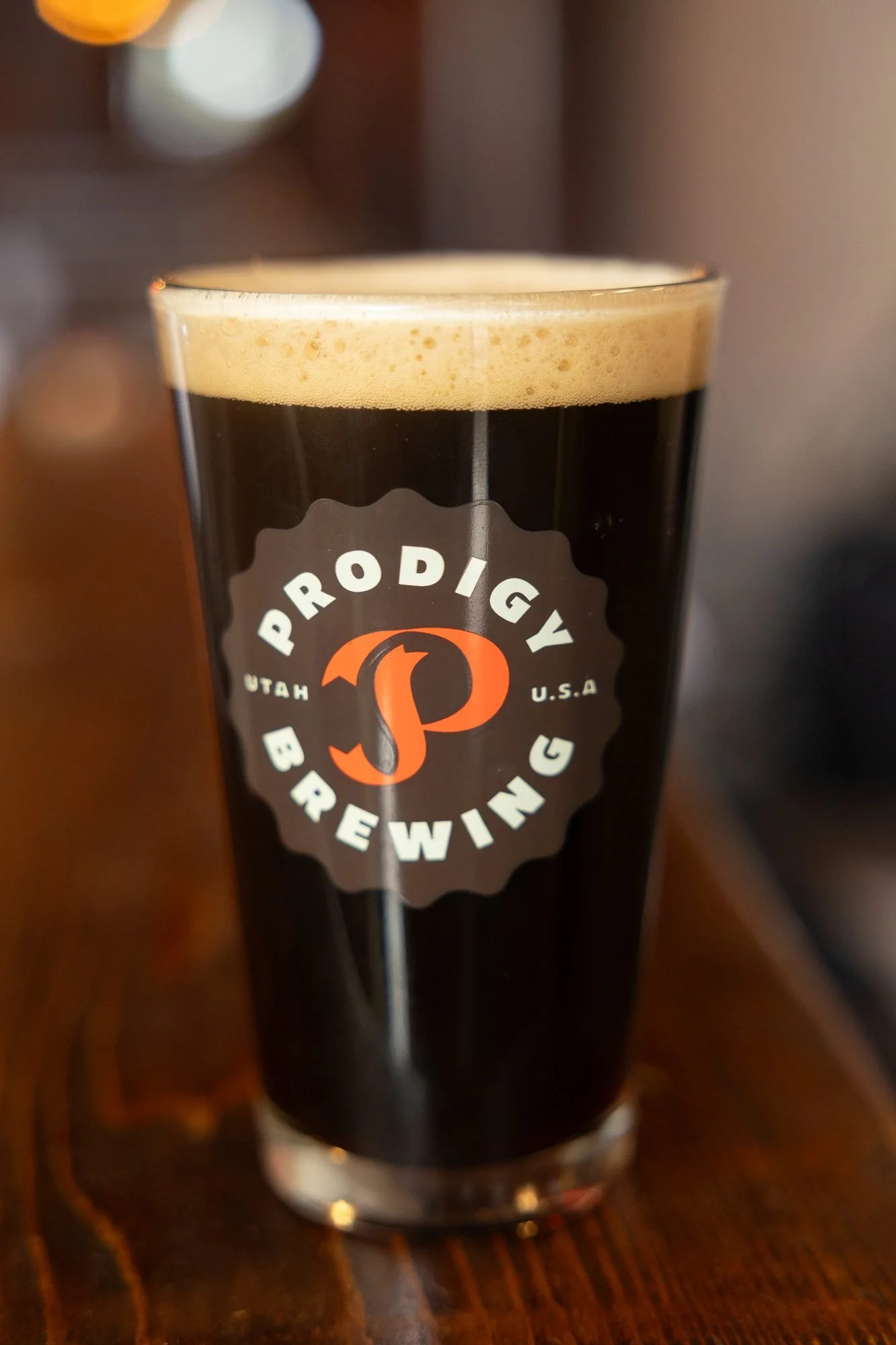 A Prodigy Brewing pint glass of dark beer with foam on a wooden bar