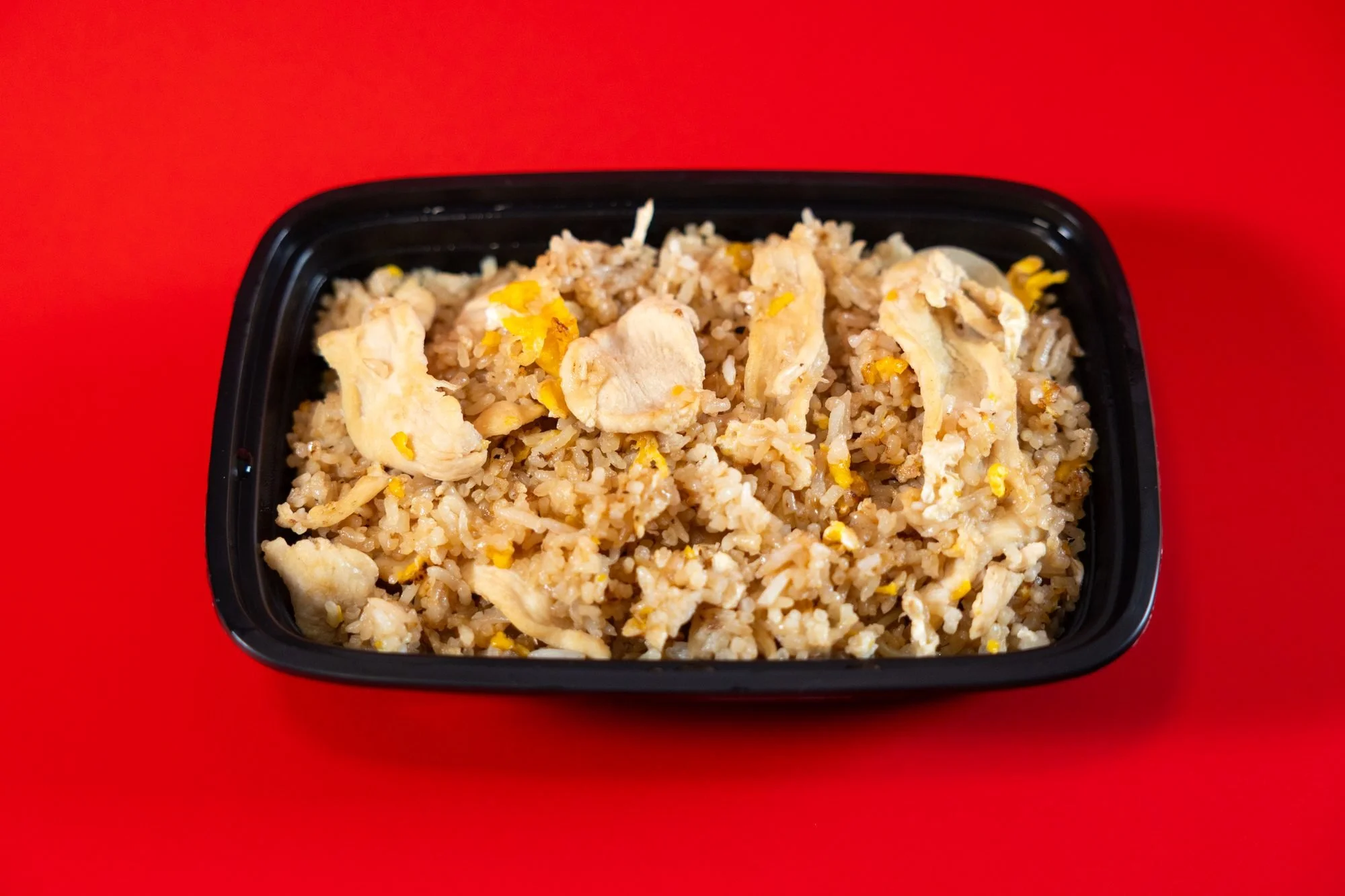 A black takeout container of chicken fried rice on a bright red background