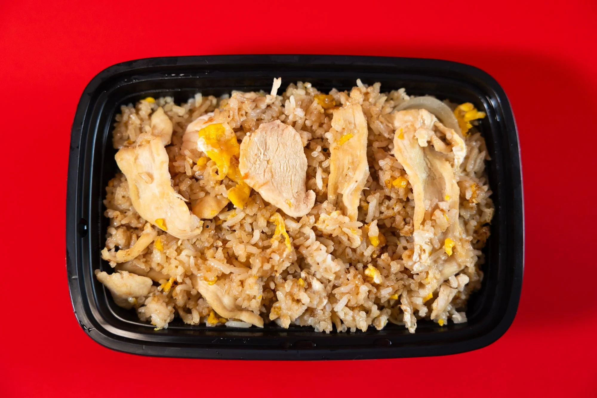 A black takeout container of fried rice with sliced chicken on a red background, shot closer