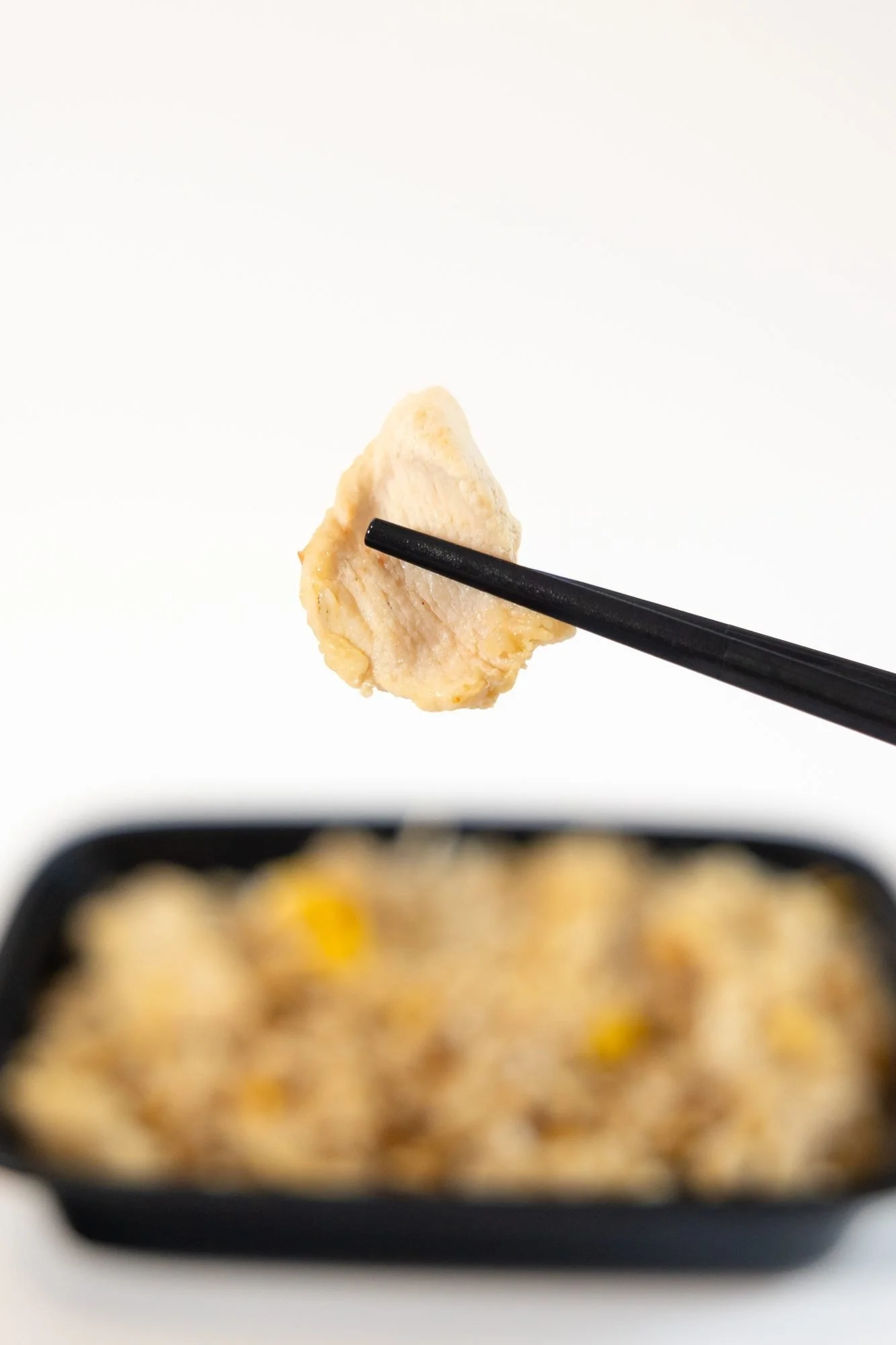 Chopsticks lift a piece of chicken above a blurred container of fried rice on a white background