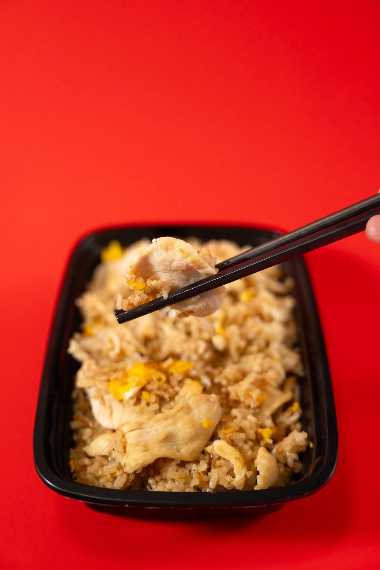 Chopsticks lift a piece of chicken from a black container of fried rice on a red background