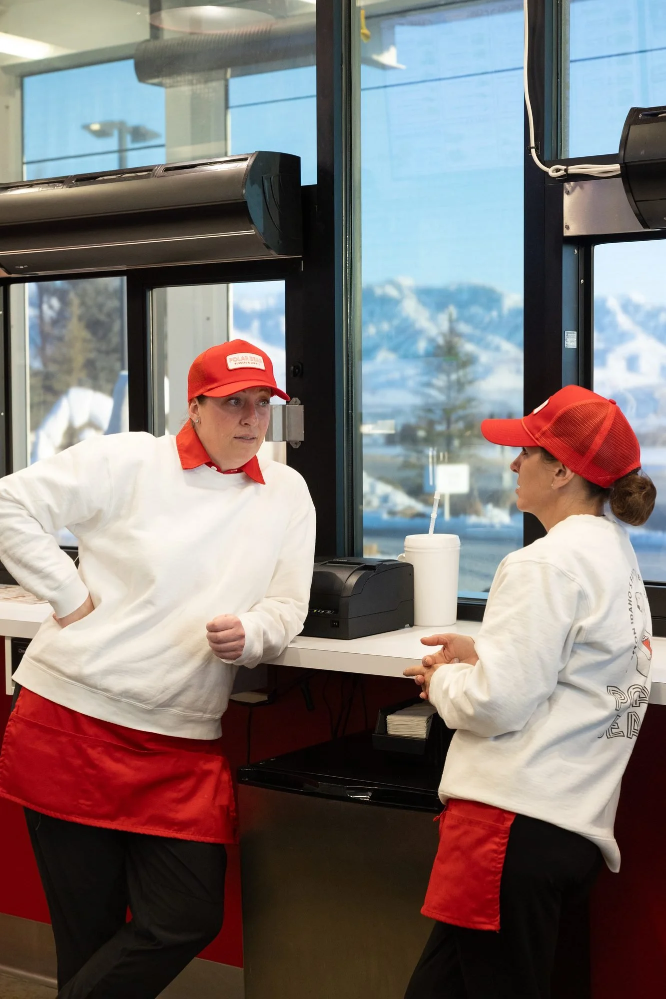 Two Polar Bear employees in white shirts and red aprons chat at the counter by a large window