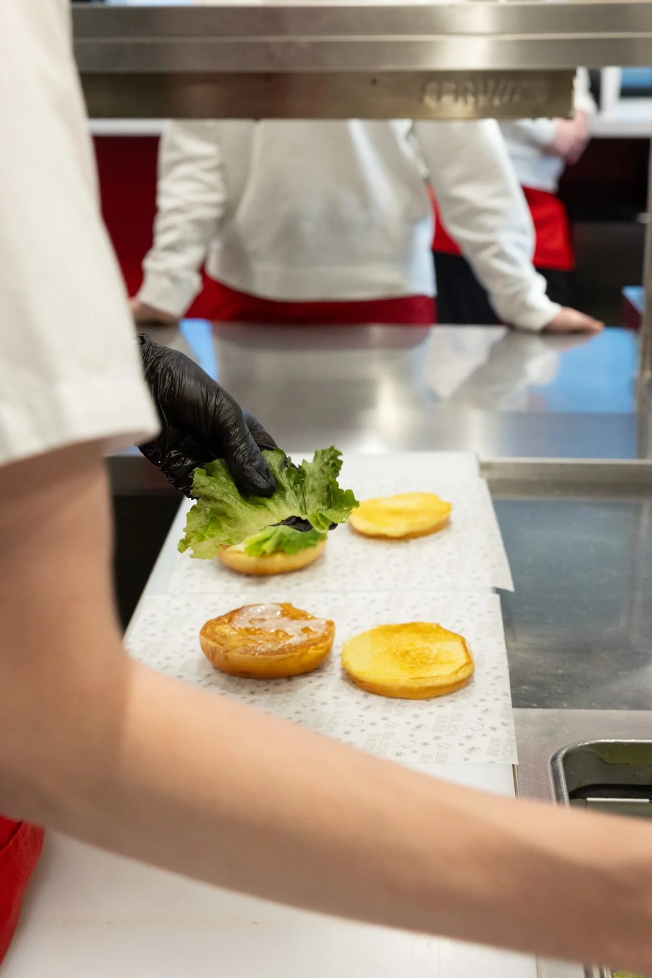 A gloved hand places lettuce on a burger bun on a steel kitchen counter