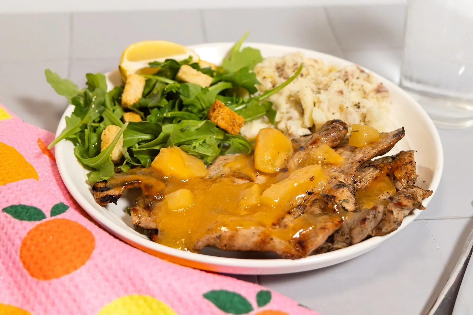 A plate of chicken with peach sauce, arugula and rice beside a fruit-print towel and water glass