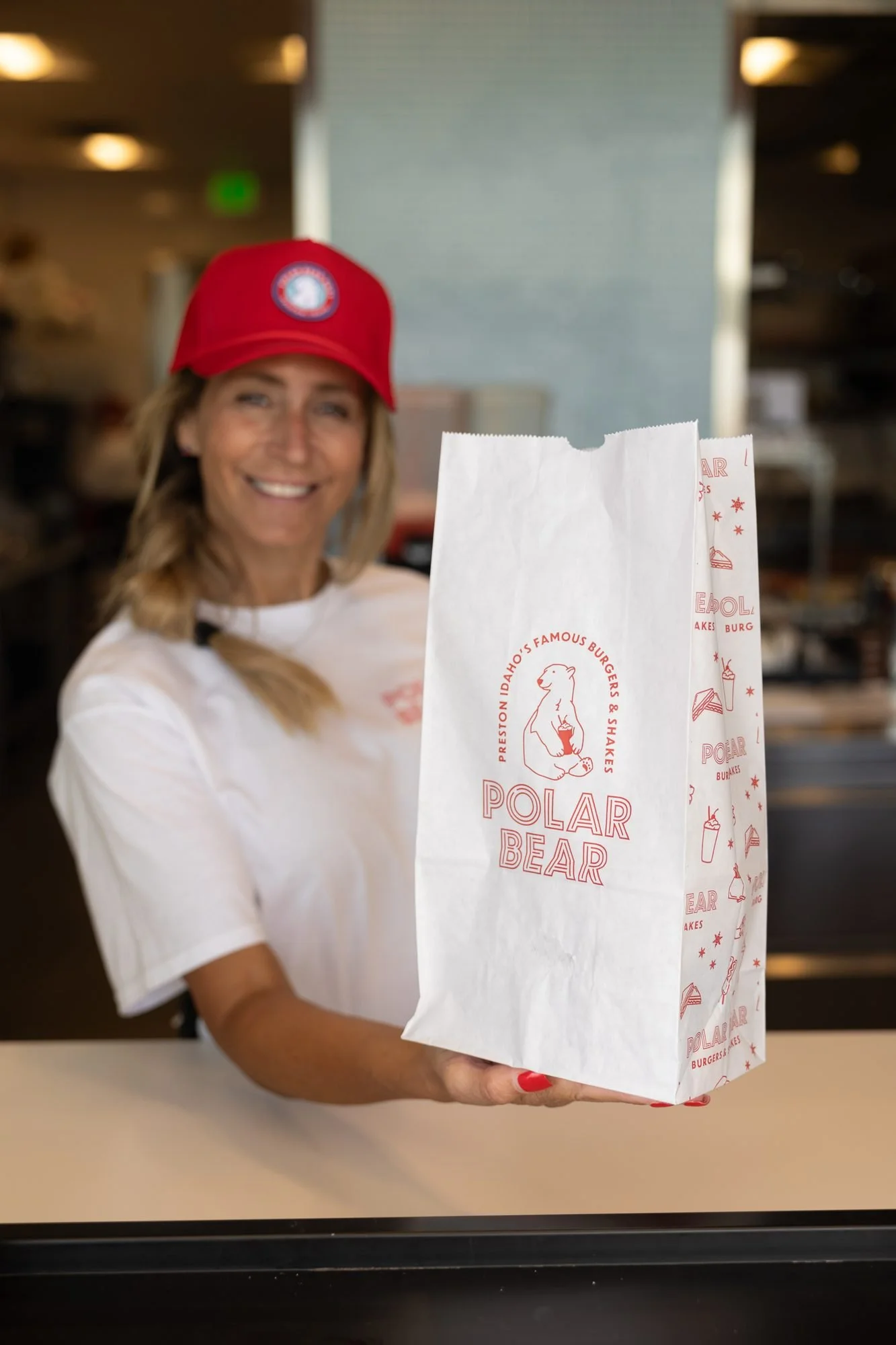 A smiling Polar Bear employee in a red cap handing a paper takeout bag through the window