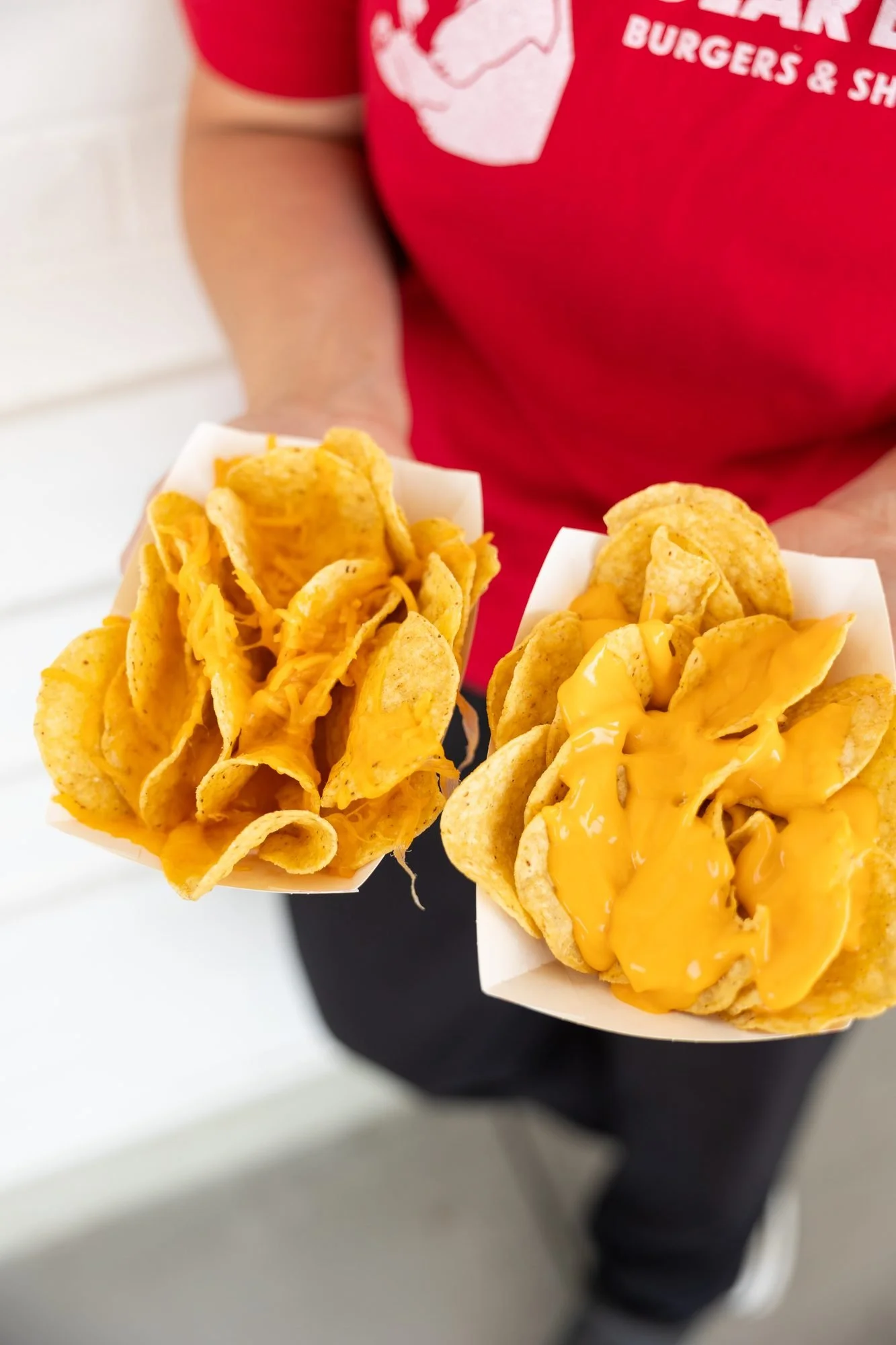 A person in a red Polar Bear shirt holding up two paper boats of cheese-covered tortilla chips