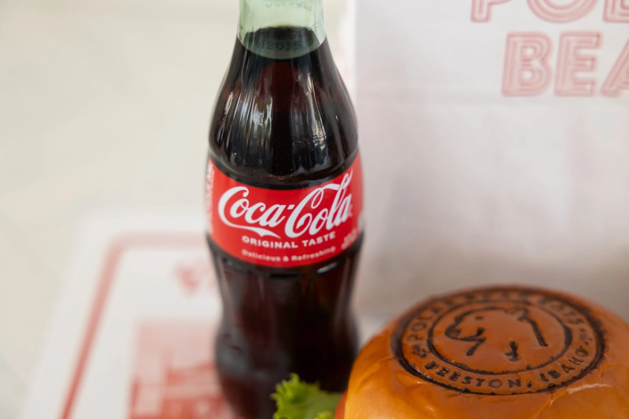A glass bottle of Coca-Cola beside a Polar Bear burger with a branded bun on a tray liner