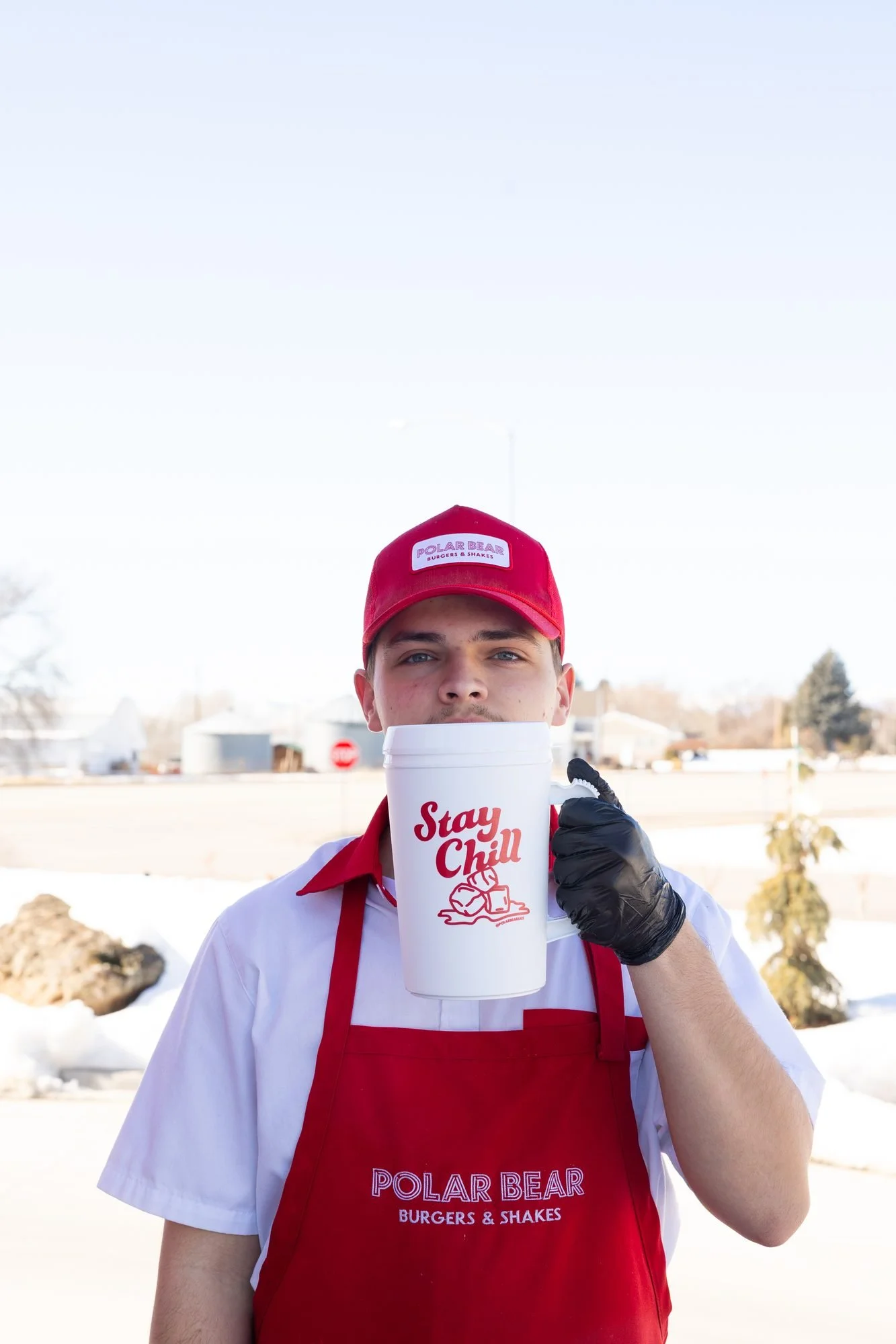 A Polar Bear employee in a red cap and apron holding a Stay Chill mug outdoors in the snow