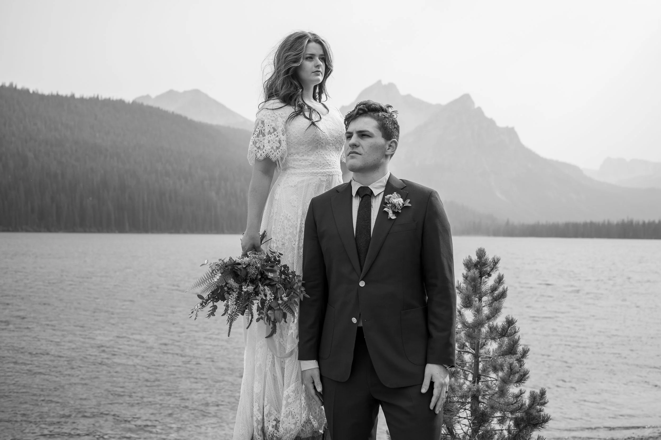 Black and white photo of a couple standing outdoors near a lake with mountains in the background. The woman wears a lace dress and holds a bouquet, while the man is in a suit with a boutonniere.