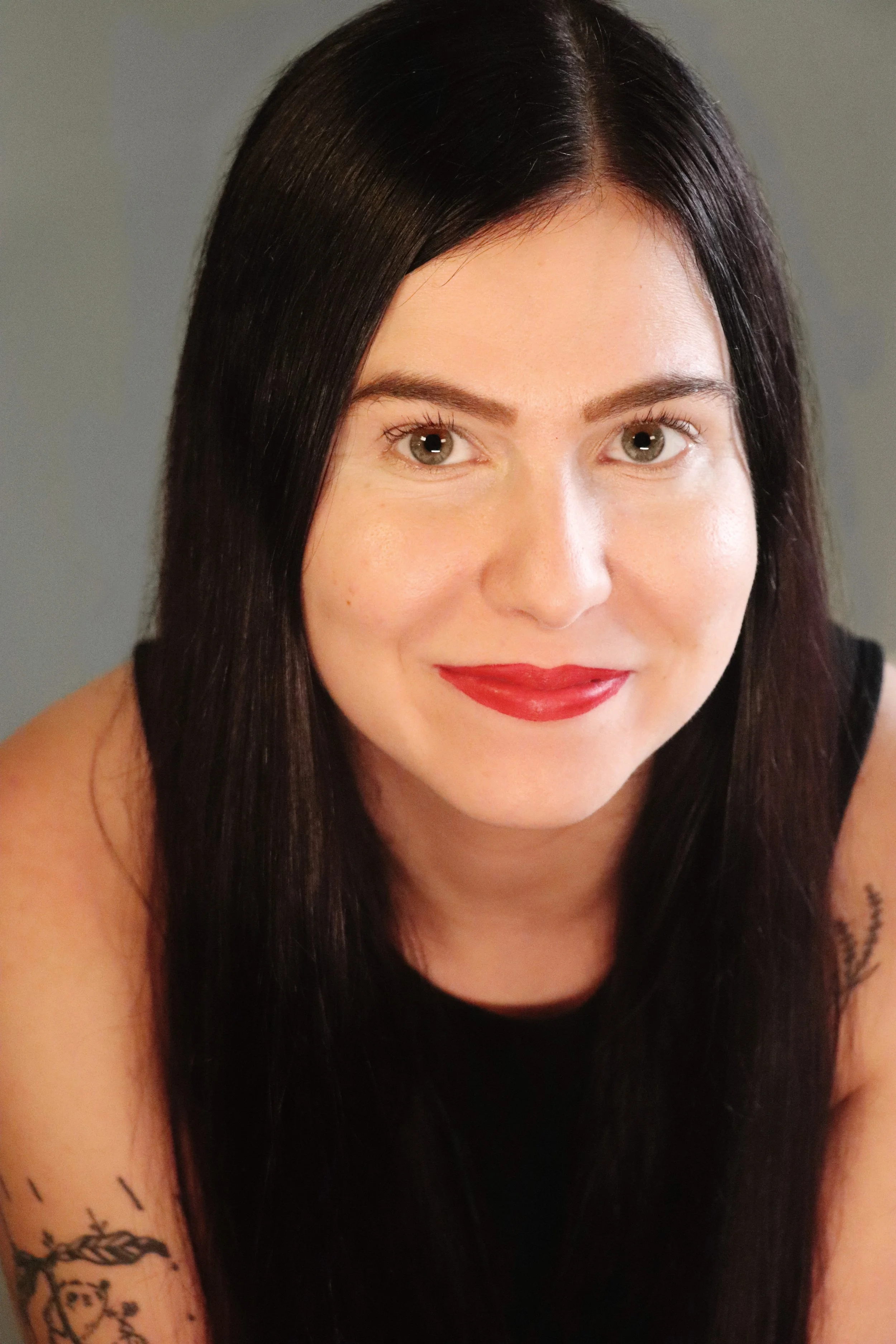 Close-up of a female-presenting person with long dark hair, fair skin, red lipstick, and a tattoo on her arm.