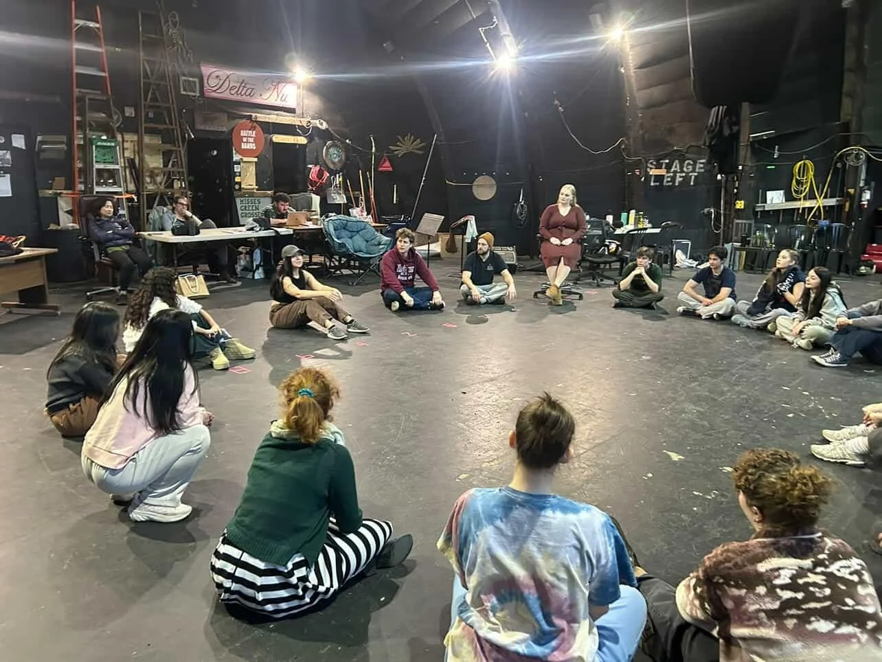 Group of people sitting in a circle on the floor inside a dimly lit rehearsal space.