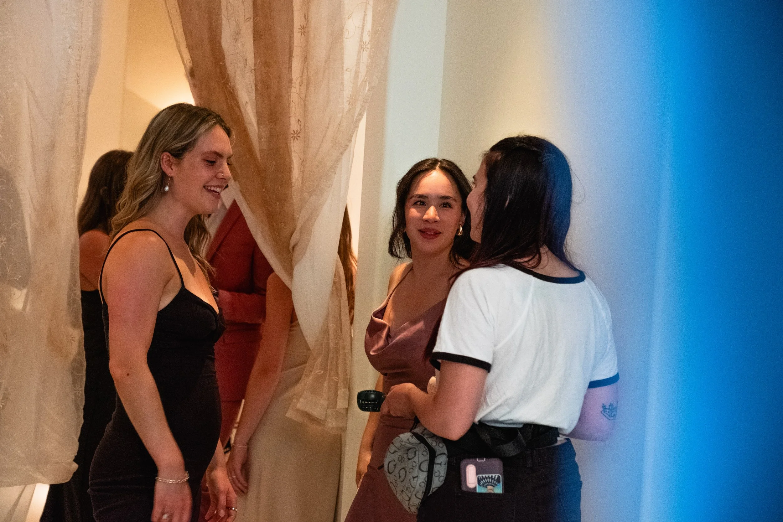 Group of three female-presenting people engaged in conversation, standing near a beige curtain in an indoor setting.