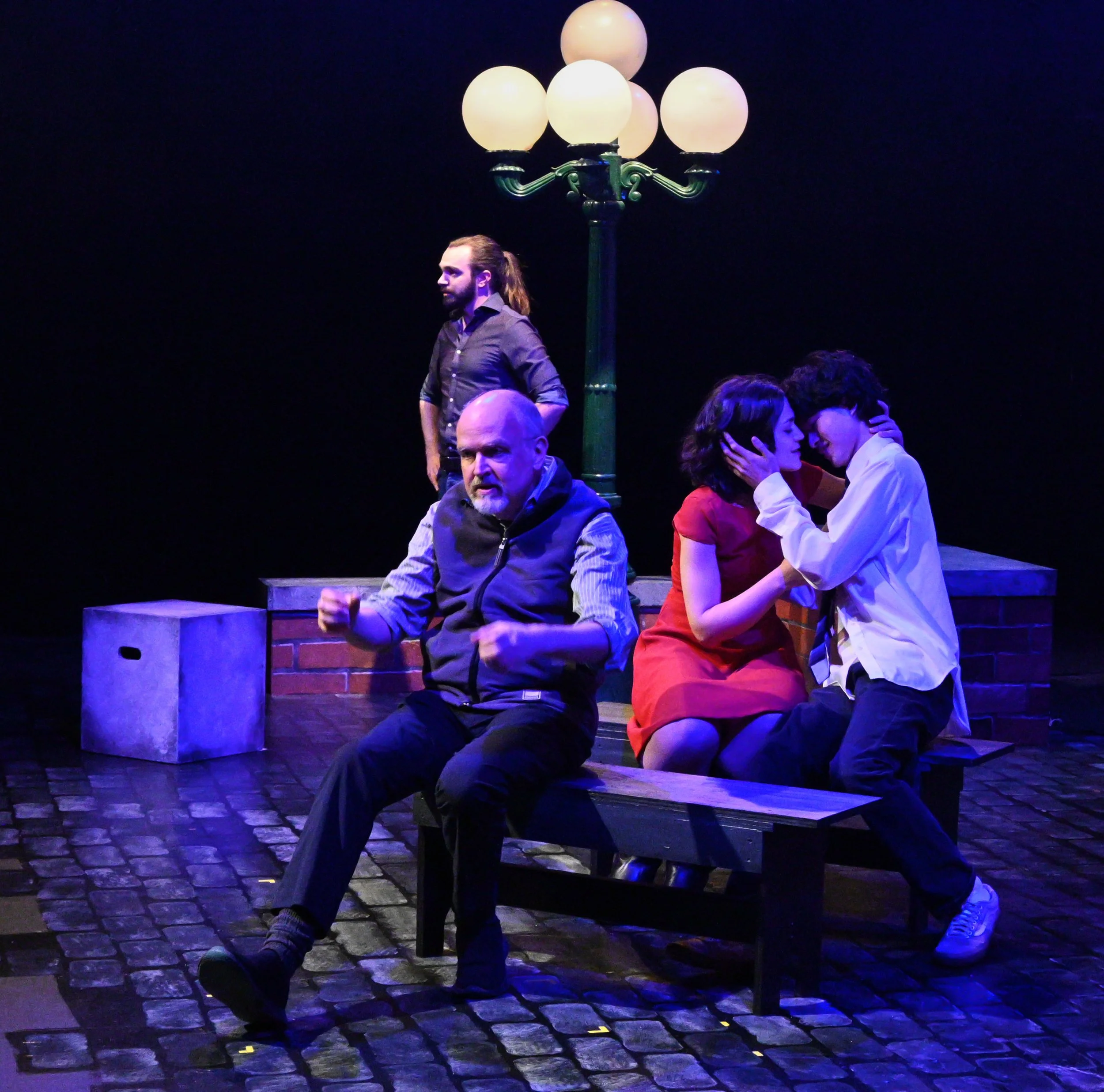 Stage scene with a man with long hair and beard standing near a vintage streetlamp, next to two people sitting on a bench kissing, against a dark background.