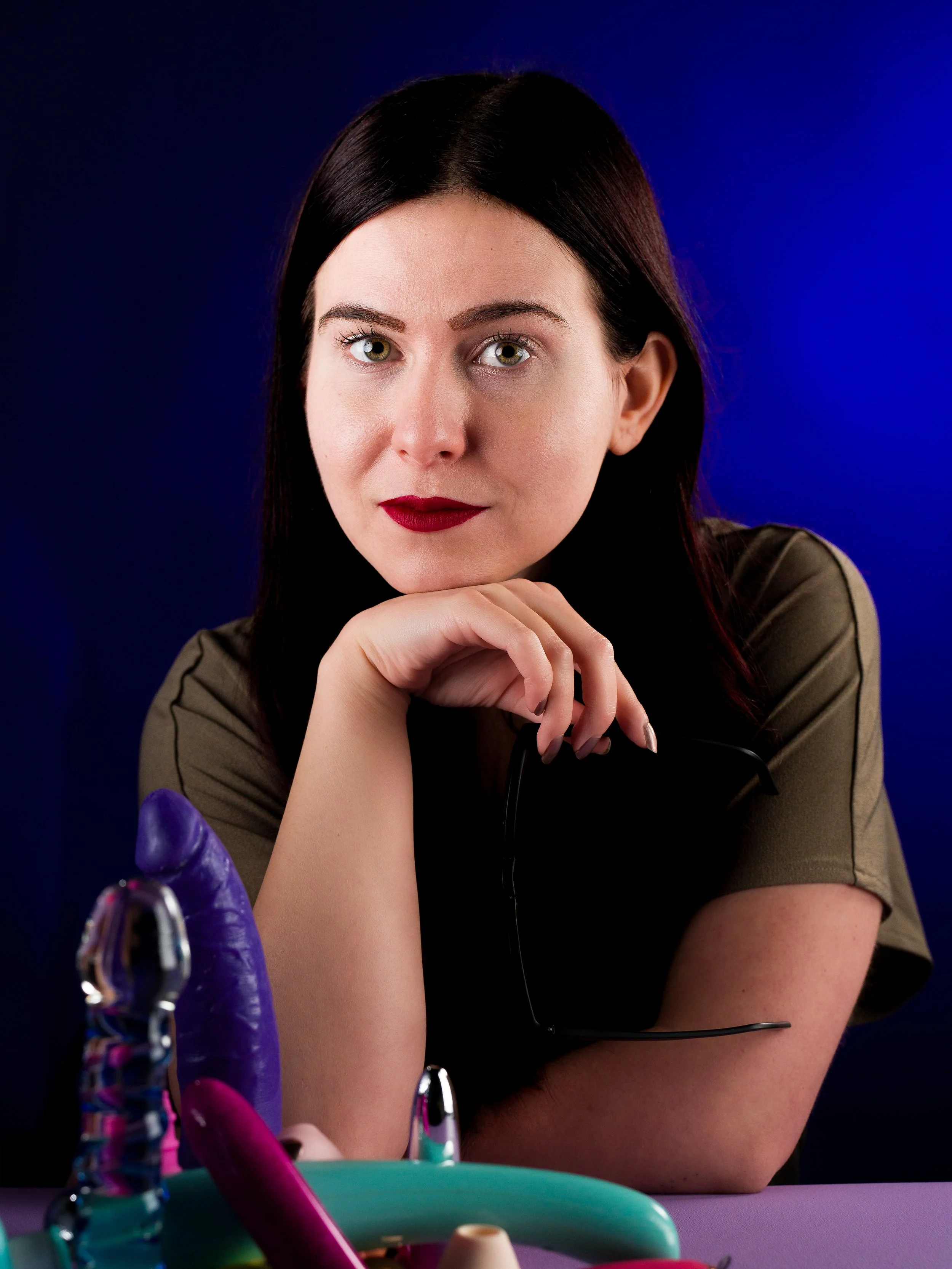 A woman with dark hair and green eyes resting her chin on her hand, looking at the camera. She is wearing makeup with red lipstick. In front of her, there are colourful toys on a purple surface. The background has a dark and blue gradient.