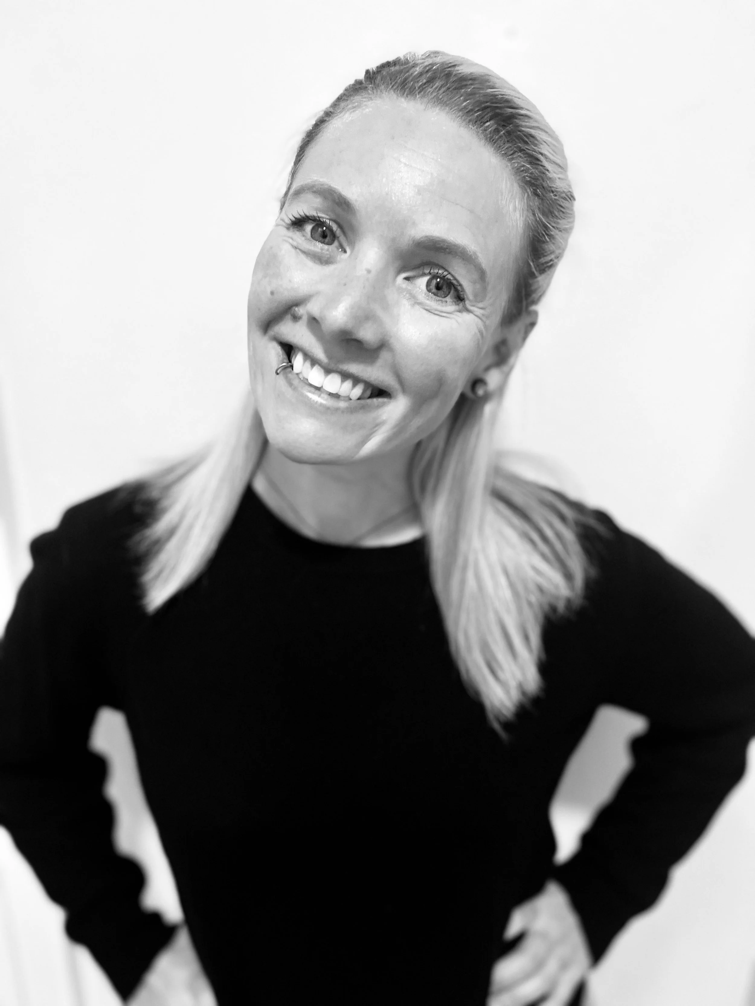 Black and white photograph of a smiling woman with long, straight hair, wearing a dark top, with her hands on her hips, standing against a plain background.